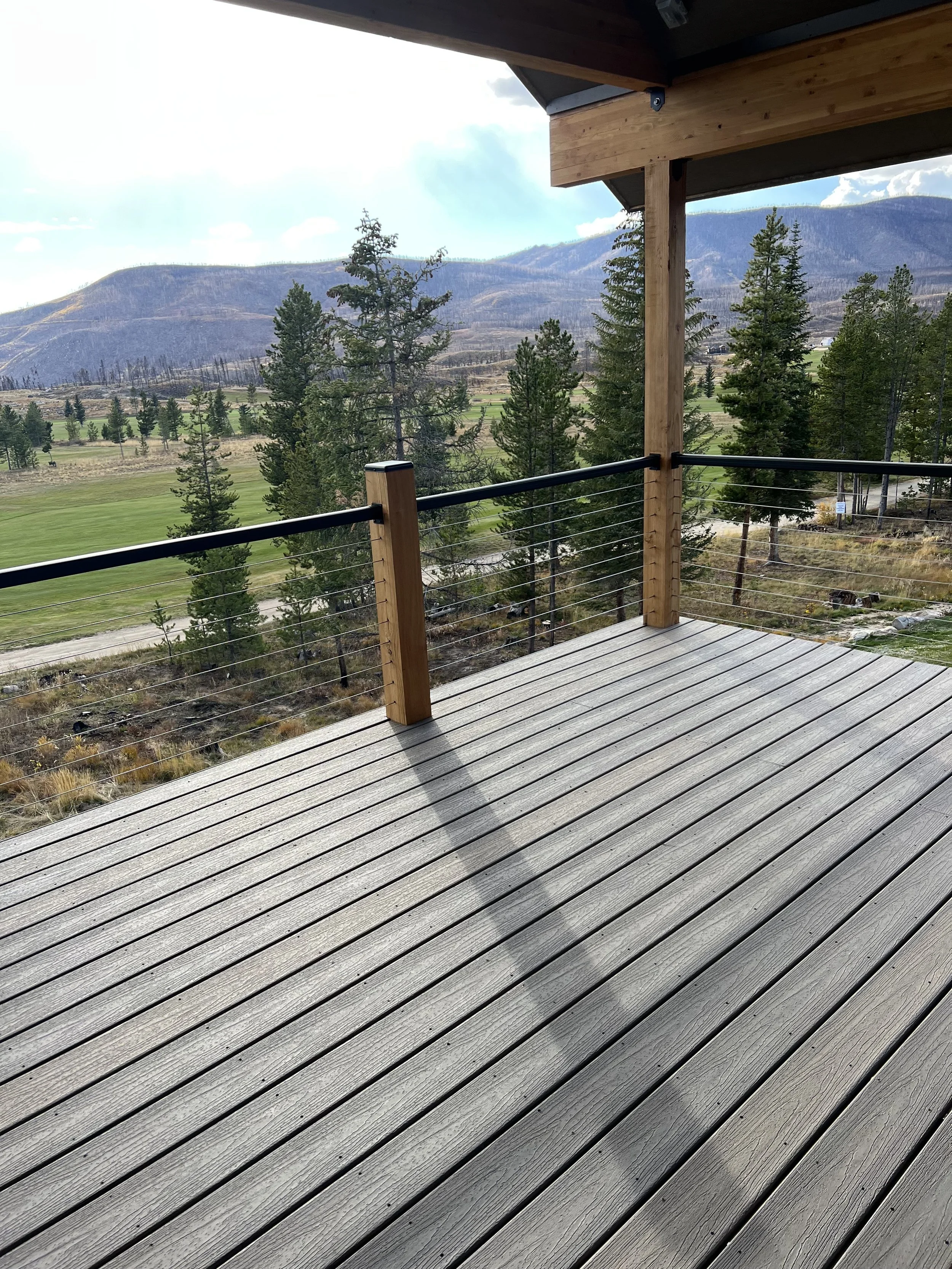 A wooden deck with metal railing overlooking a landscape of trees, grassy fields, hills, and mountains under a partly cloudy sky.