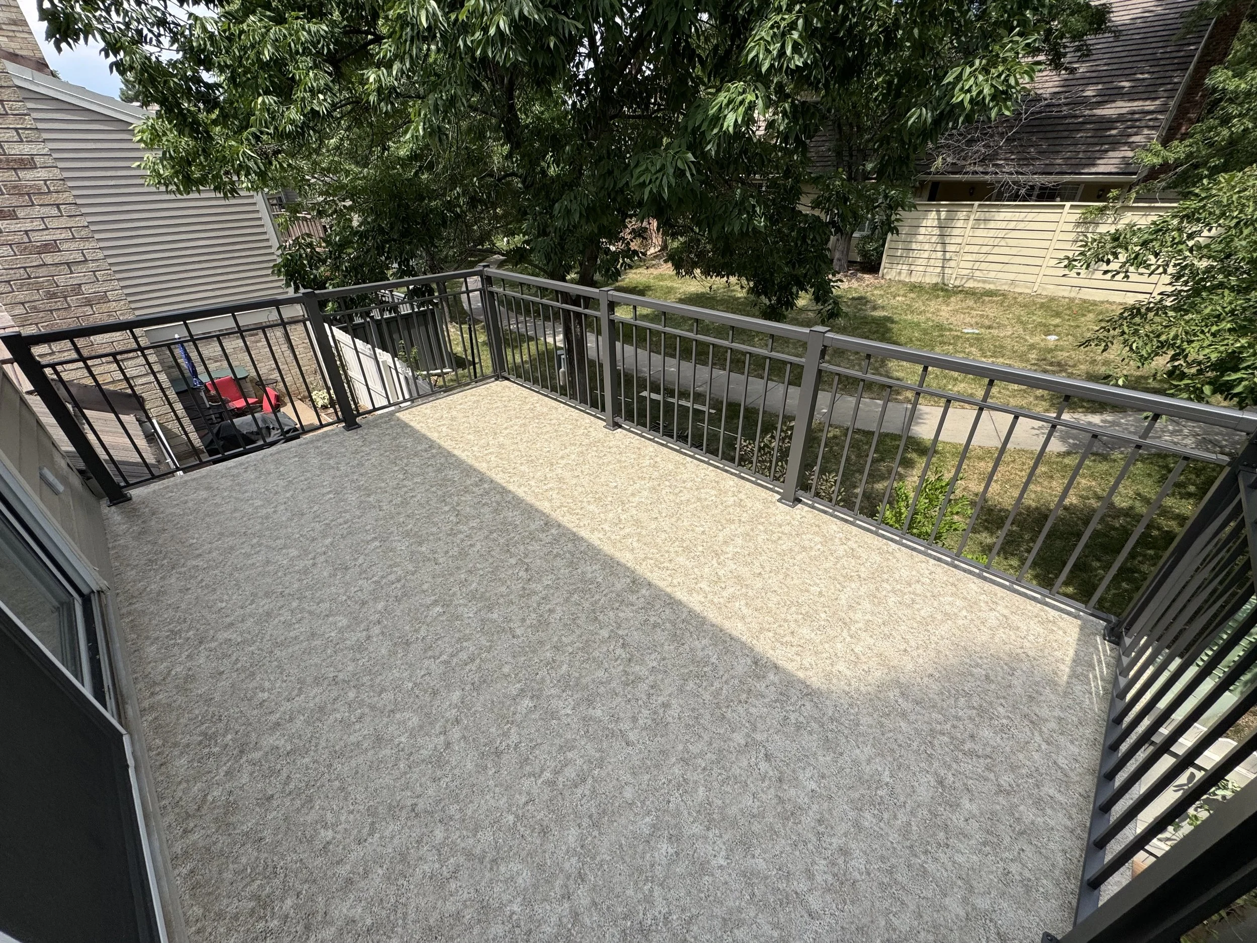 A view of a small balcony with beige railing, overlooking a backyard with grass, trees, and a sidewalk, with neighboring houses visible in the background.