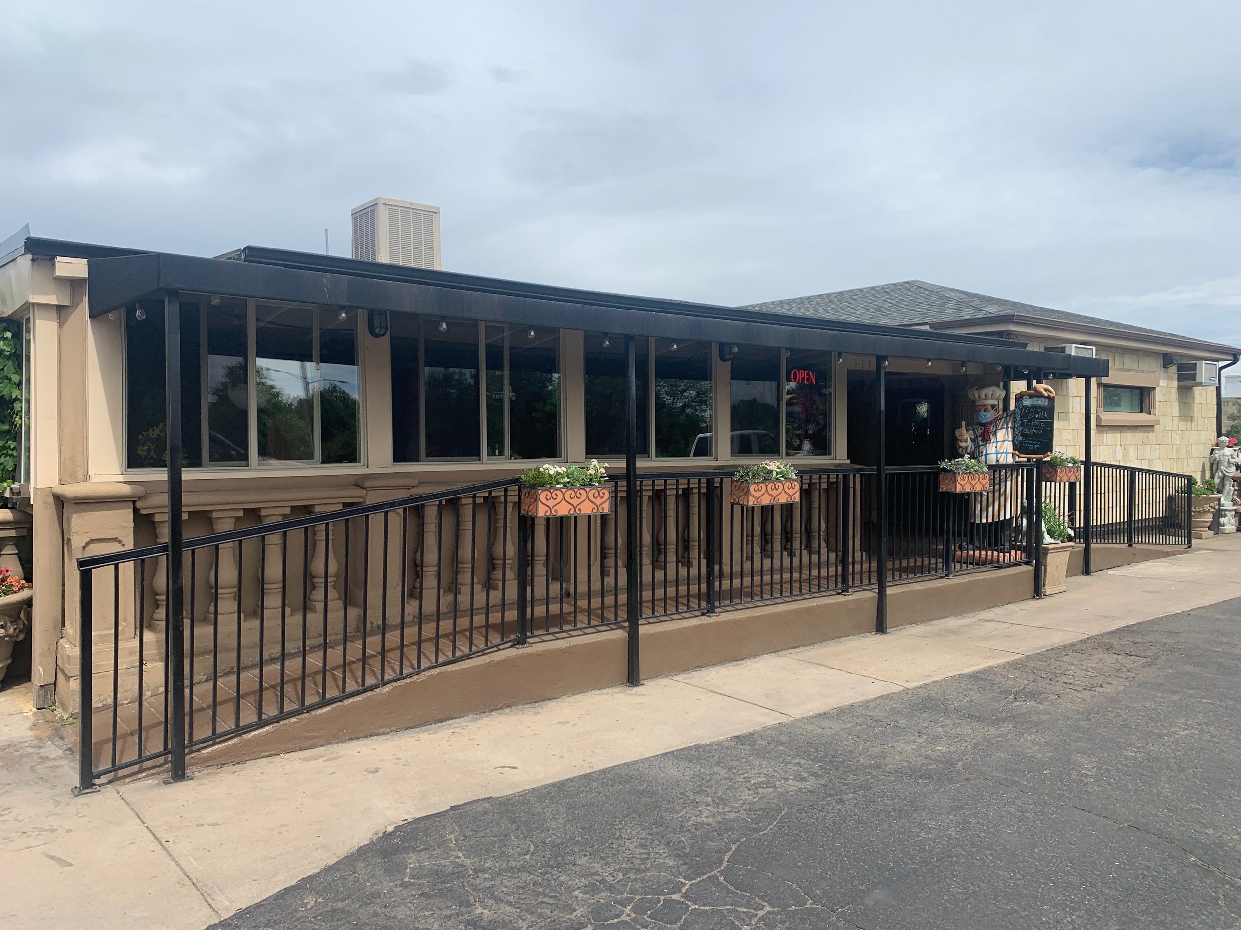 Exterior of a small restaurant or cafe with a covered porch, black railing, window boxes with plants, and a signboard near the entrance.