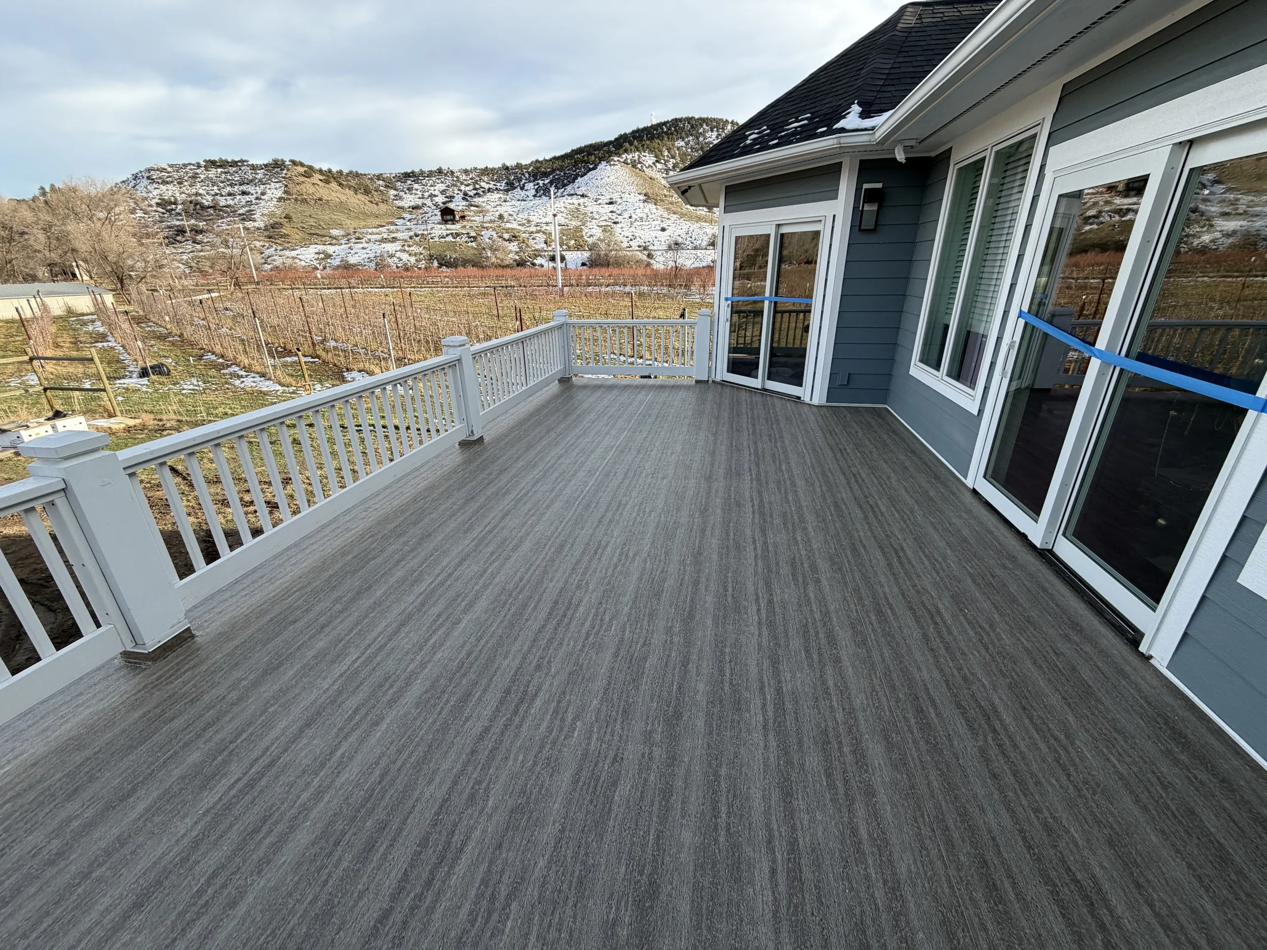 Empty outdoor balcony with gray flooring and white railing overlooking snow-capped hills and a vineyard in the background.