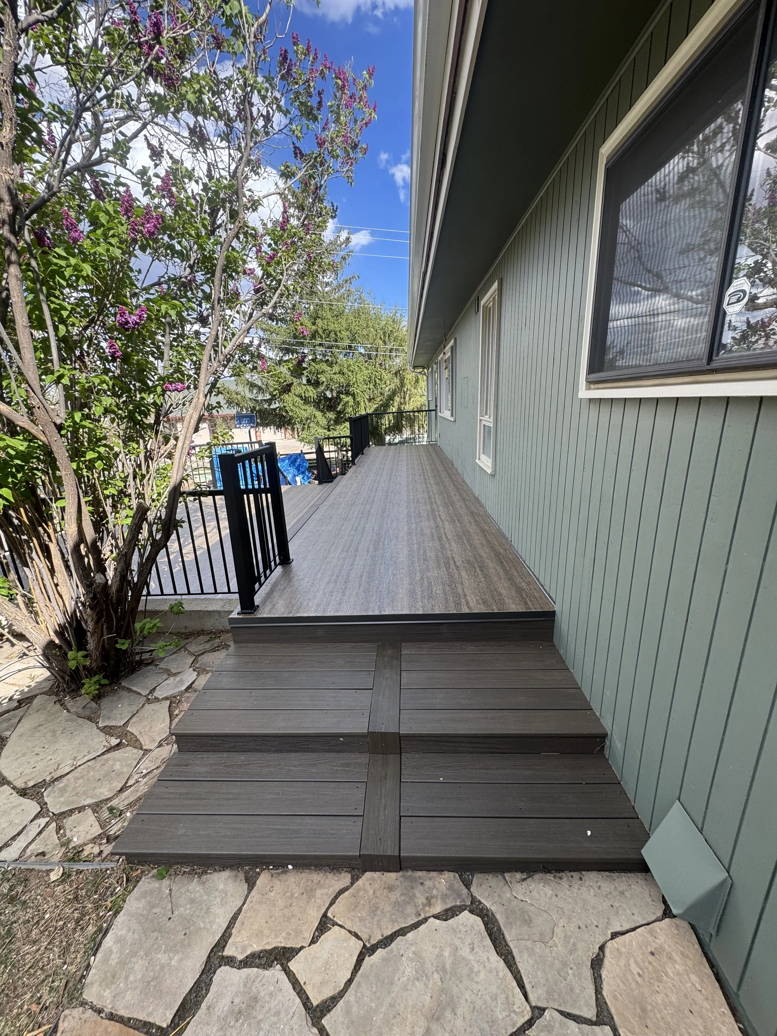 A house with a green exterior wall and a wooden deck. The deck has a small flight of three steps leading up to it, with a black metal railing on the left side. There are purple flowering trees to the left and a clear blue sky above.