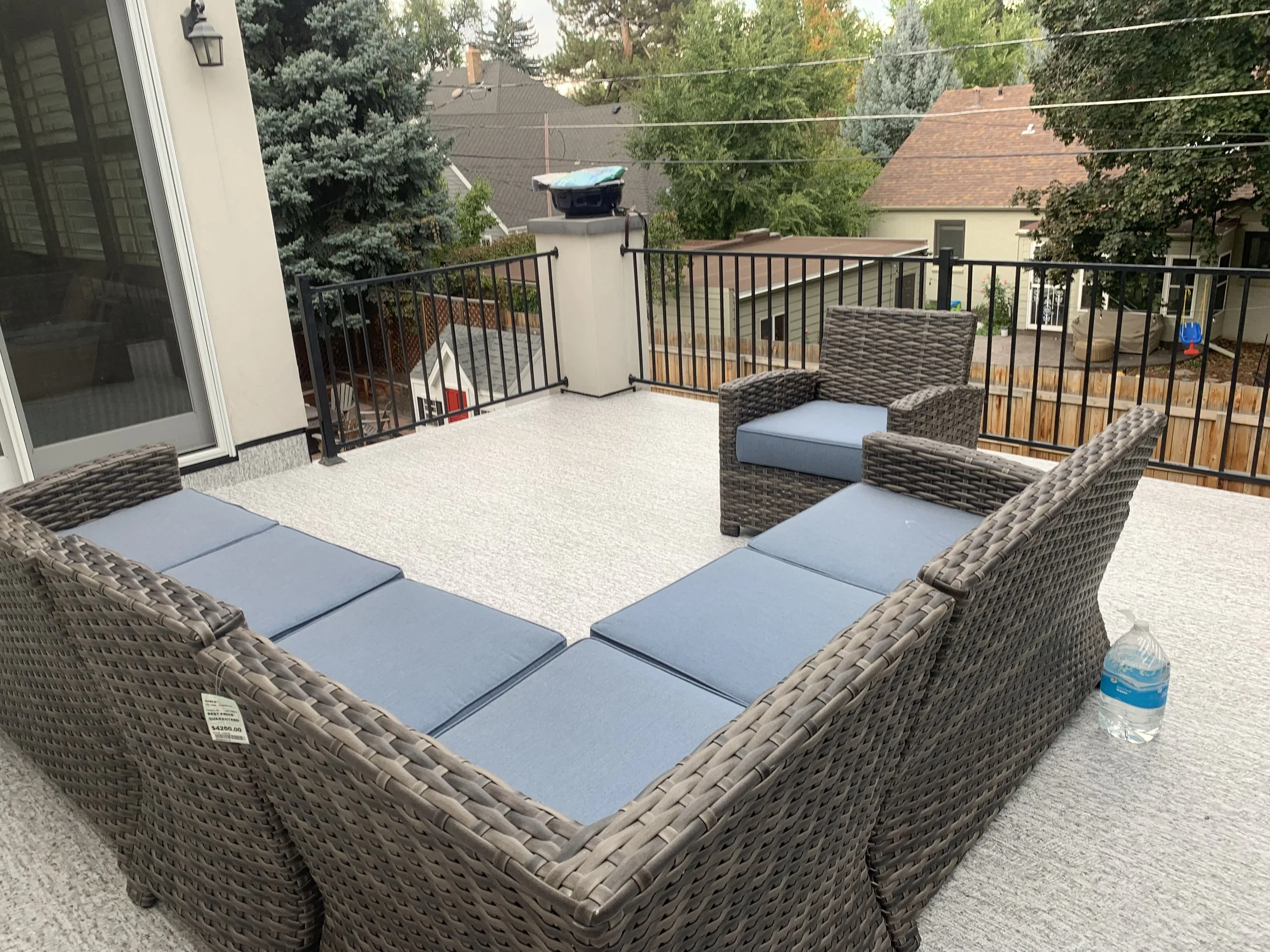 Outdoor patio with a wicker sectional sofa with blue cushions, two matching wicker armchairs with blue cushions, a plastic water bottle on the floor, and a black metal railing surrounding the deck. In the background, trees, neighboring houses, and a 