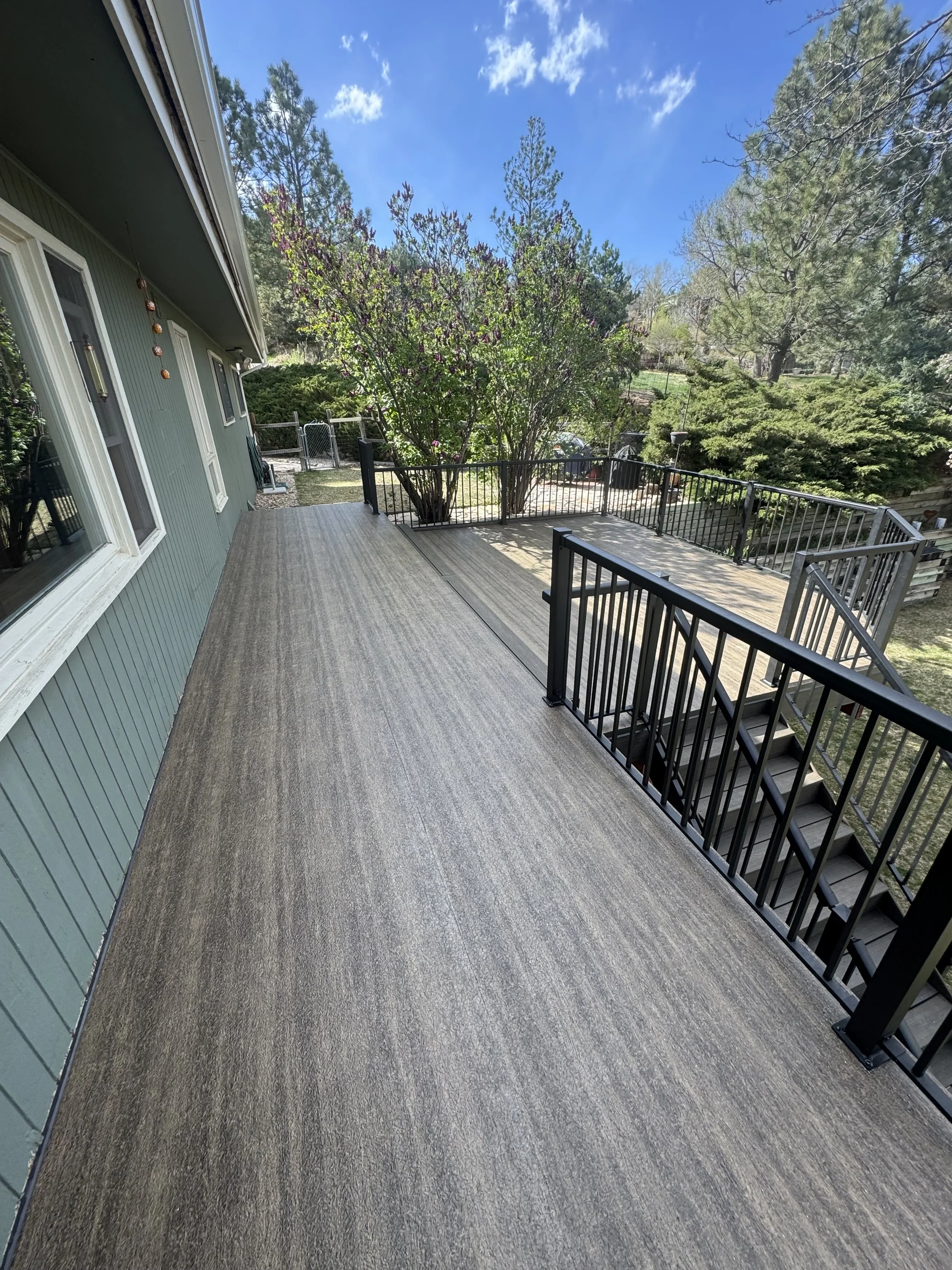 Outdoor wooden deck with black metal railing, green house with windows on the left, trim, and a lush garden with trees and bushes in the background under a partly cloudy blue sky.