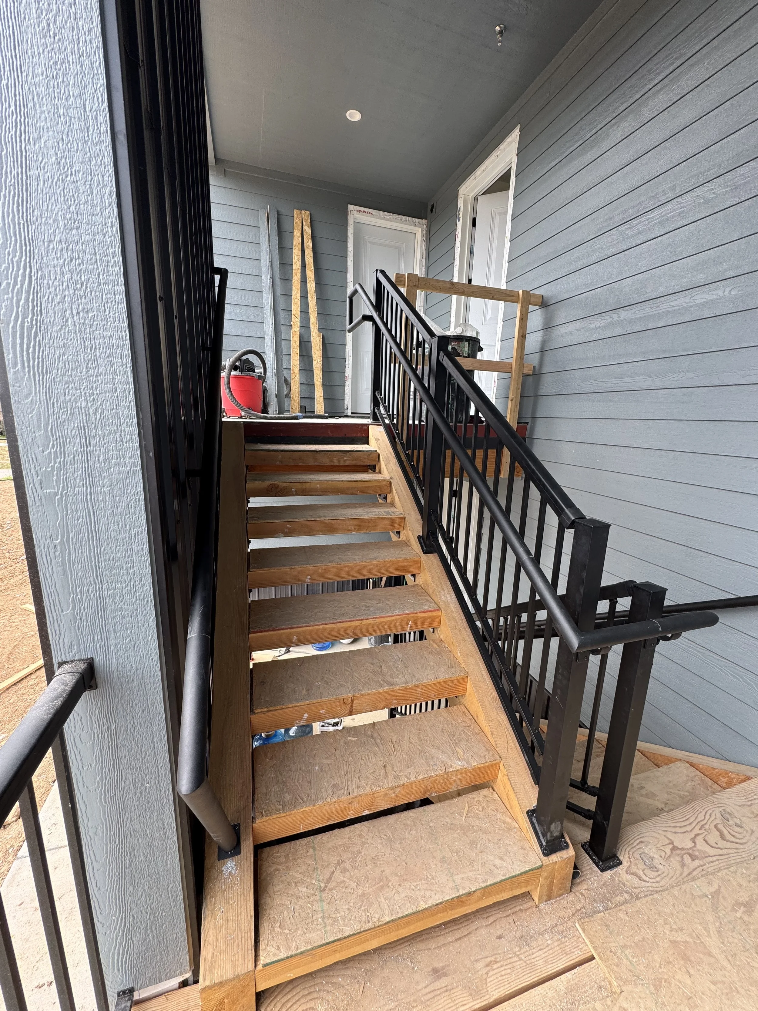 Construction site of a stairway with new wooden steps, black metal railings, gray wooden siding, and construction tools and materials in the background.