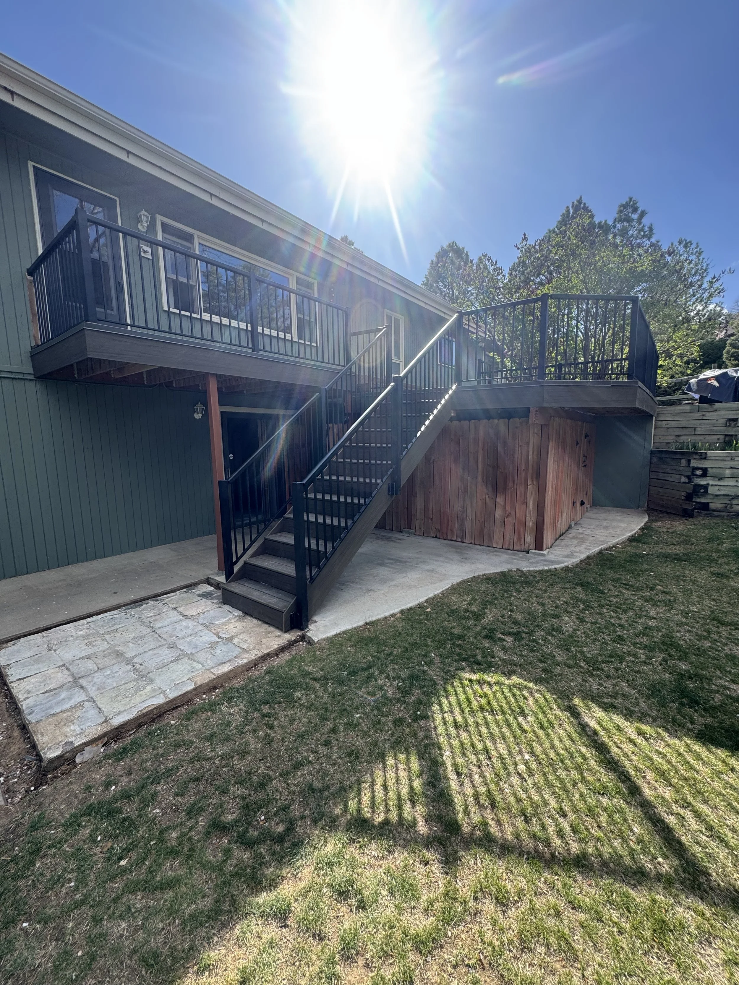 Backyard view of a house with a metal deck and staircase, with sunlight overhead and a shadow of the deck on the grassy yard.