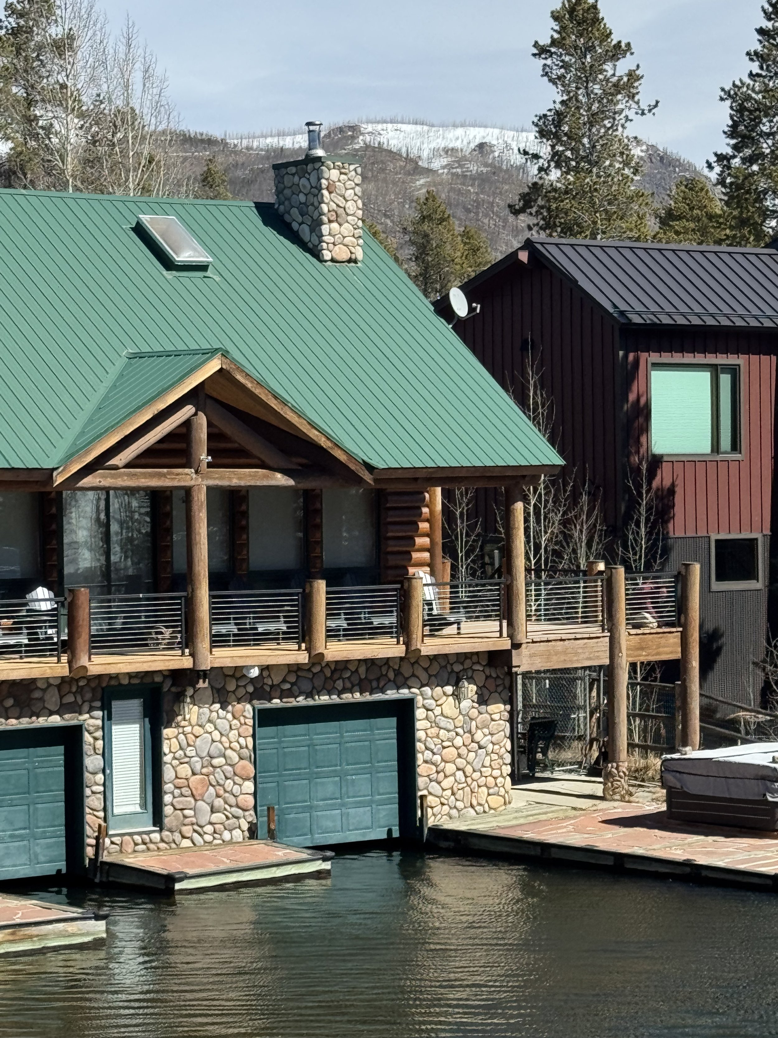 A house with a green metal roof and stone chimney next to a body of water, surrounded by trees and mountains in the background.