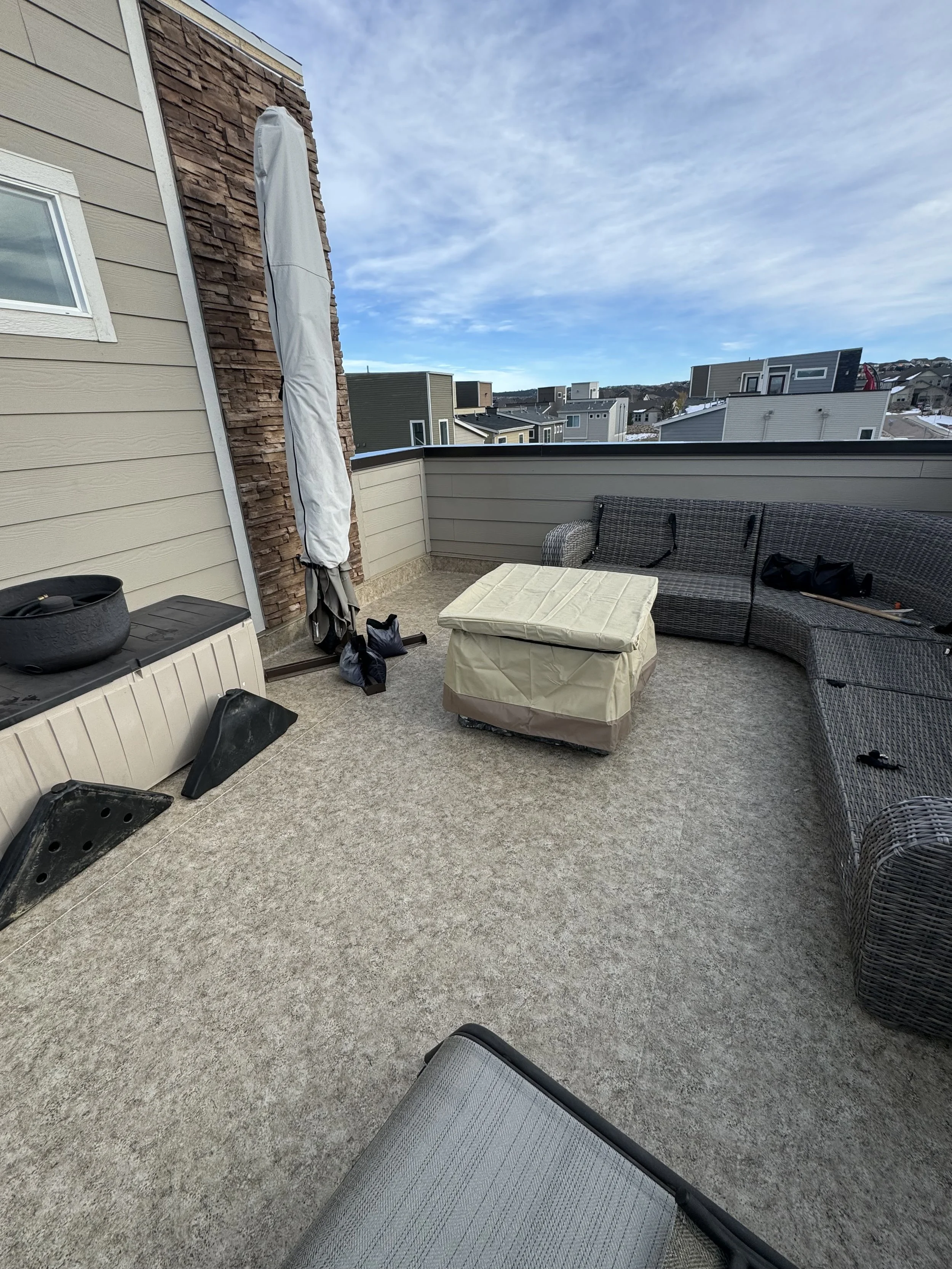 A rooftop balcony with beige and gray furniture, including a sectional wicker sofa, and a small table or ottoman. There are black tools or equipment on the sofa, a covered round fire pit or heater, and some black bags and objects on the floor near th