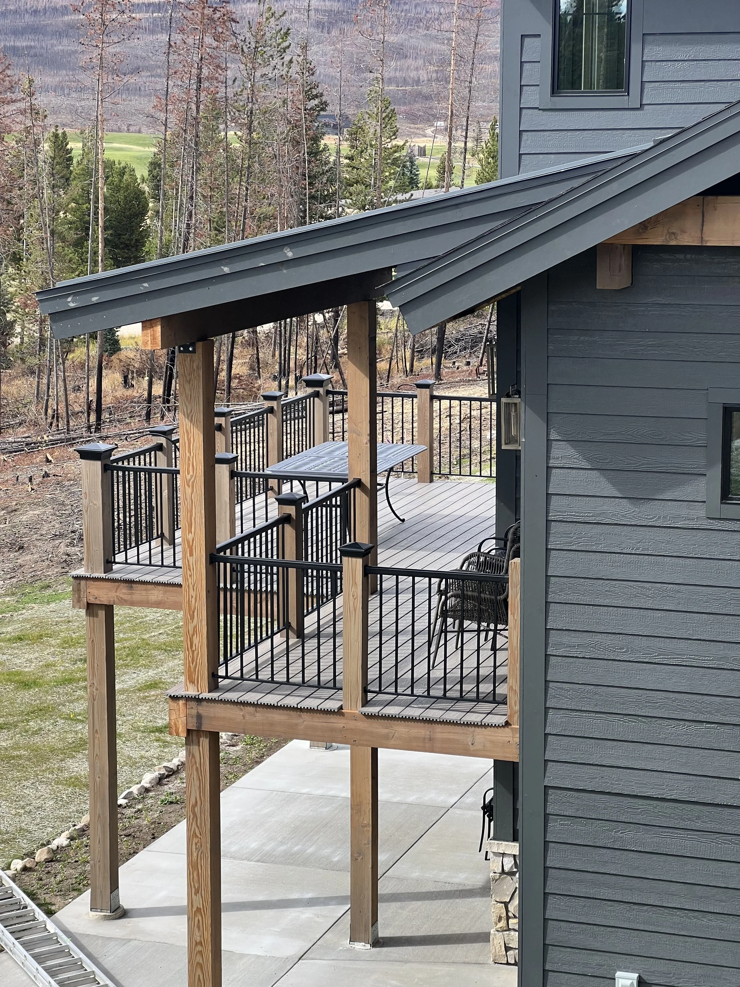 A multi-level wooden deck attached to a gray house with black metal railings, overlooking a natural landscape with trees and mountains.