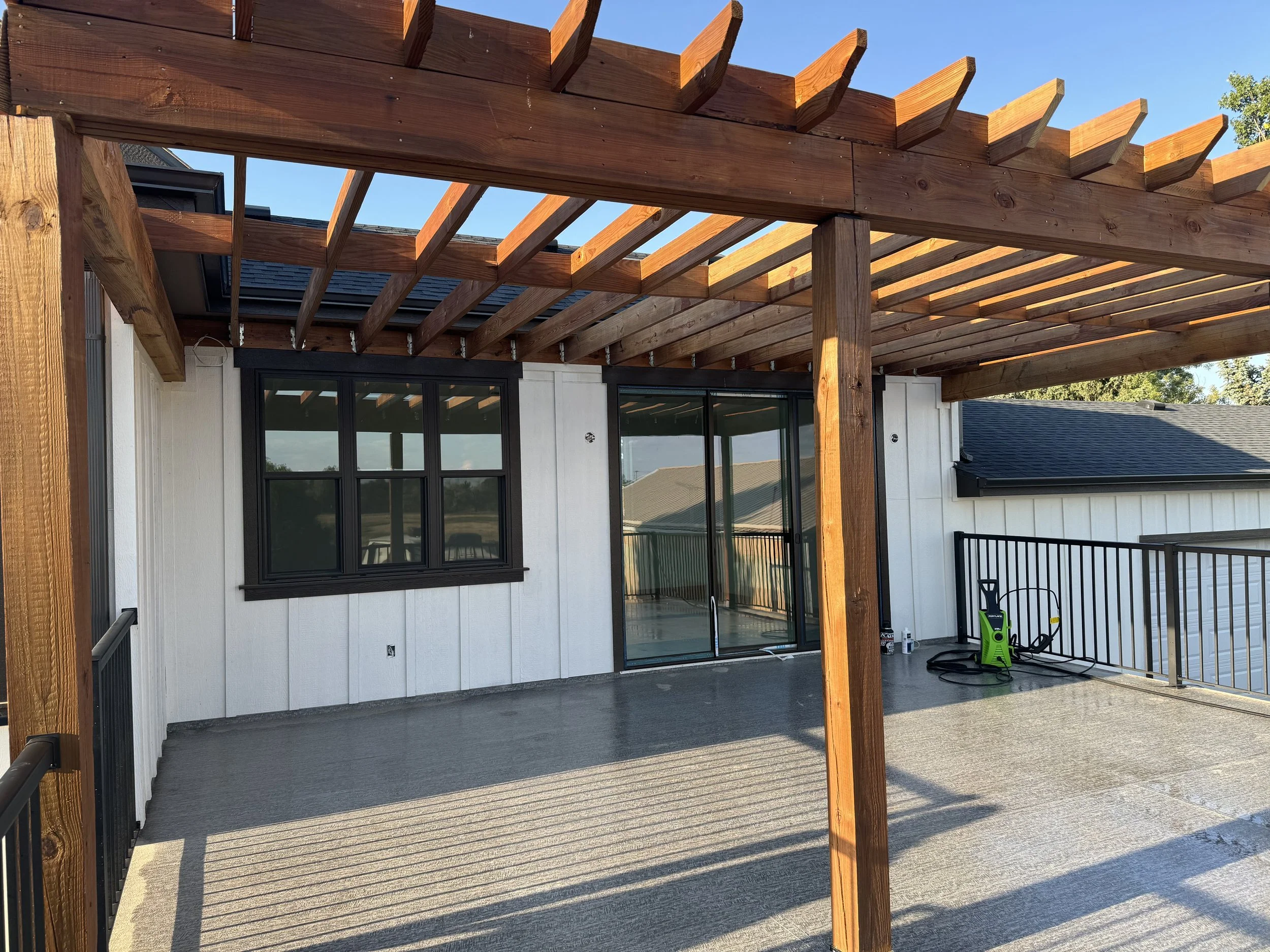 A balcony with a wooden pergola frame, sliding glass door, a window, and a spray washer on the right side. The balcony has a concrete floor and black metal railing.