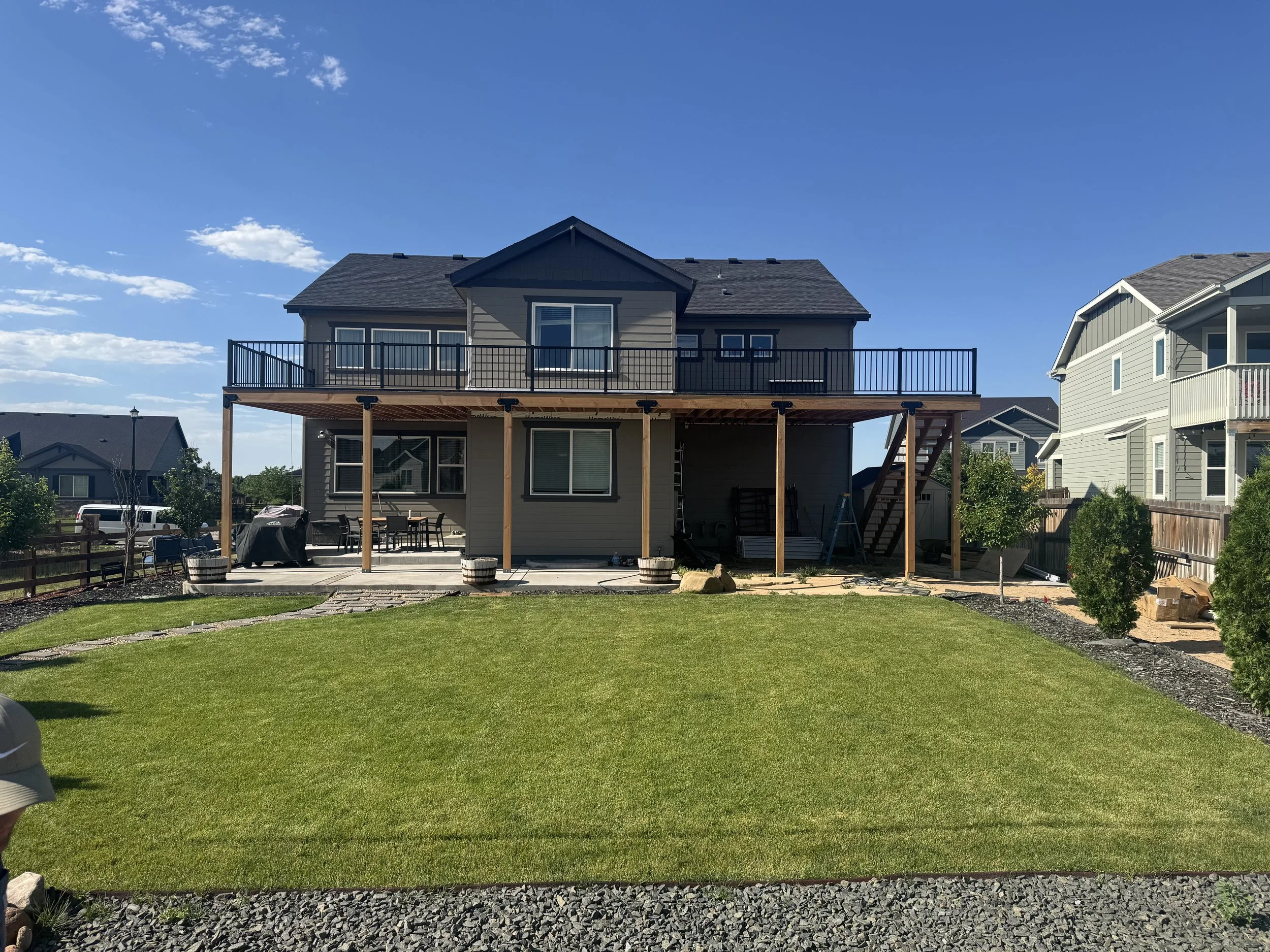 A two-story house under construction with a large back deck, surrounded by green lawn and neighboring houses.