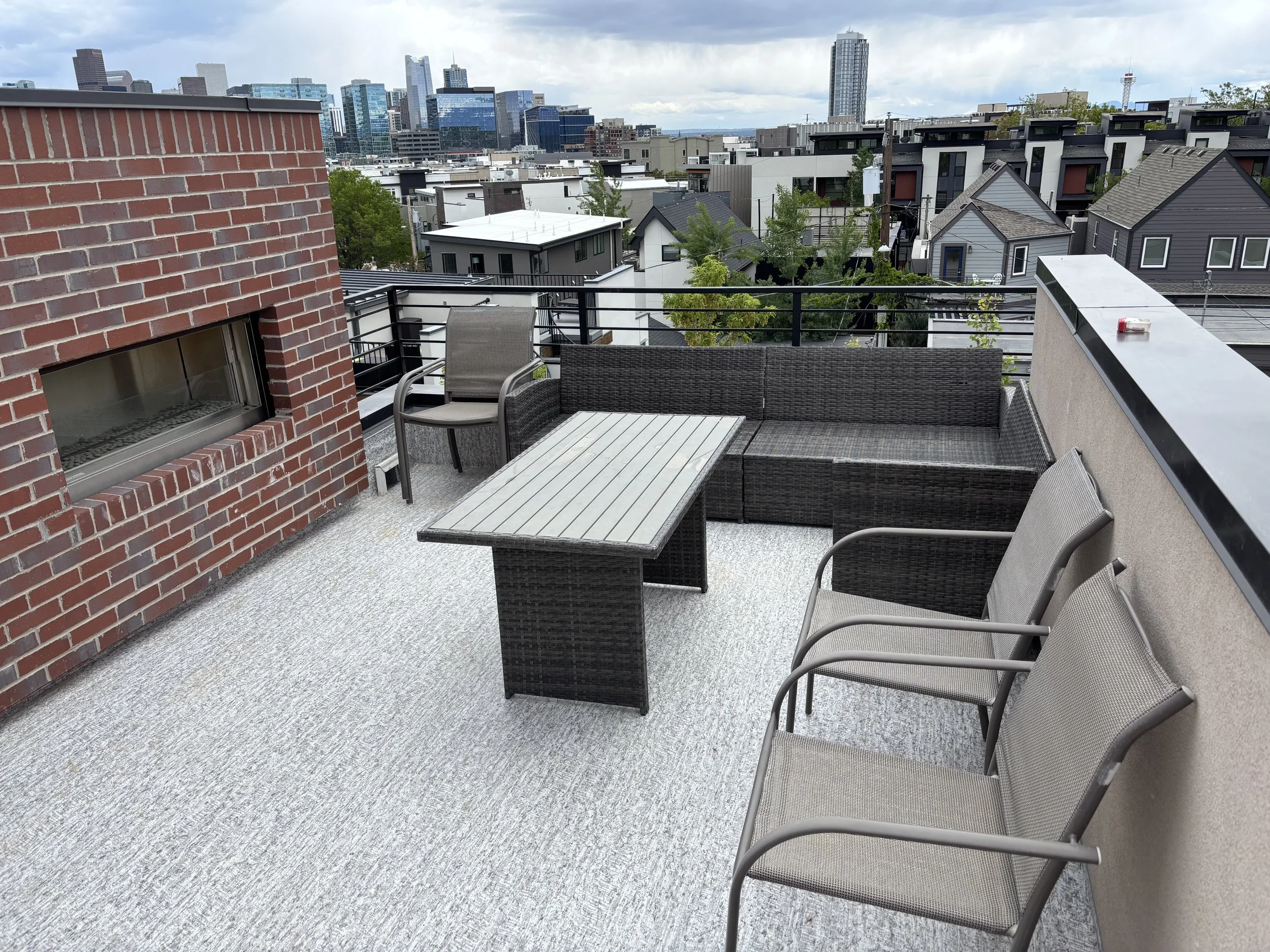 Rooftop patio with outdoor furniture including a table, chairs, and a wicker sectional sofa, overlooking a cityscape with buildings and trees under cloudy skies.