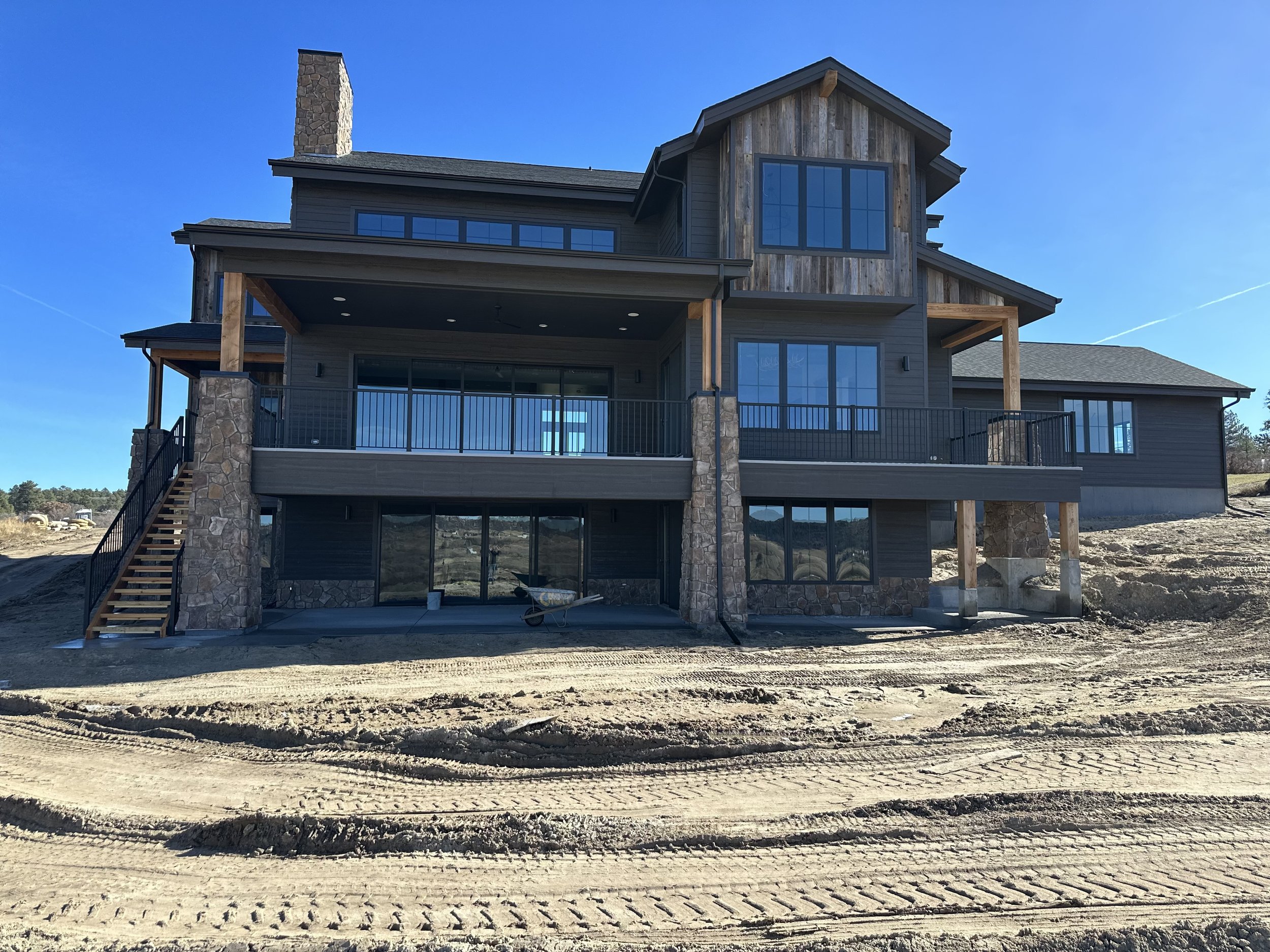 Front view of a modern multi-story house under construction with large windows, a stone chimney, and a spacious balcony, set on a dirt lot.