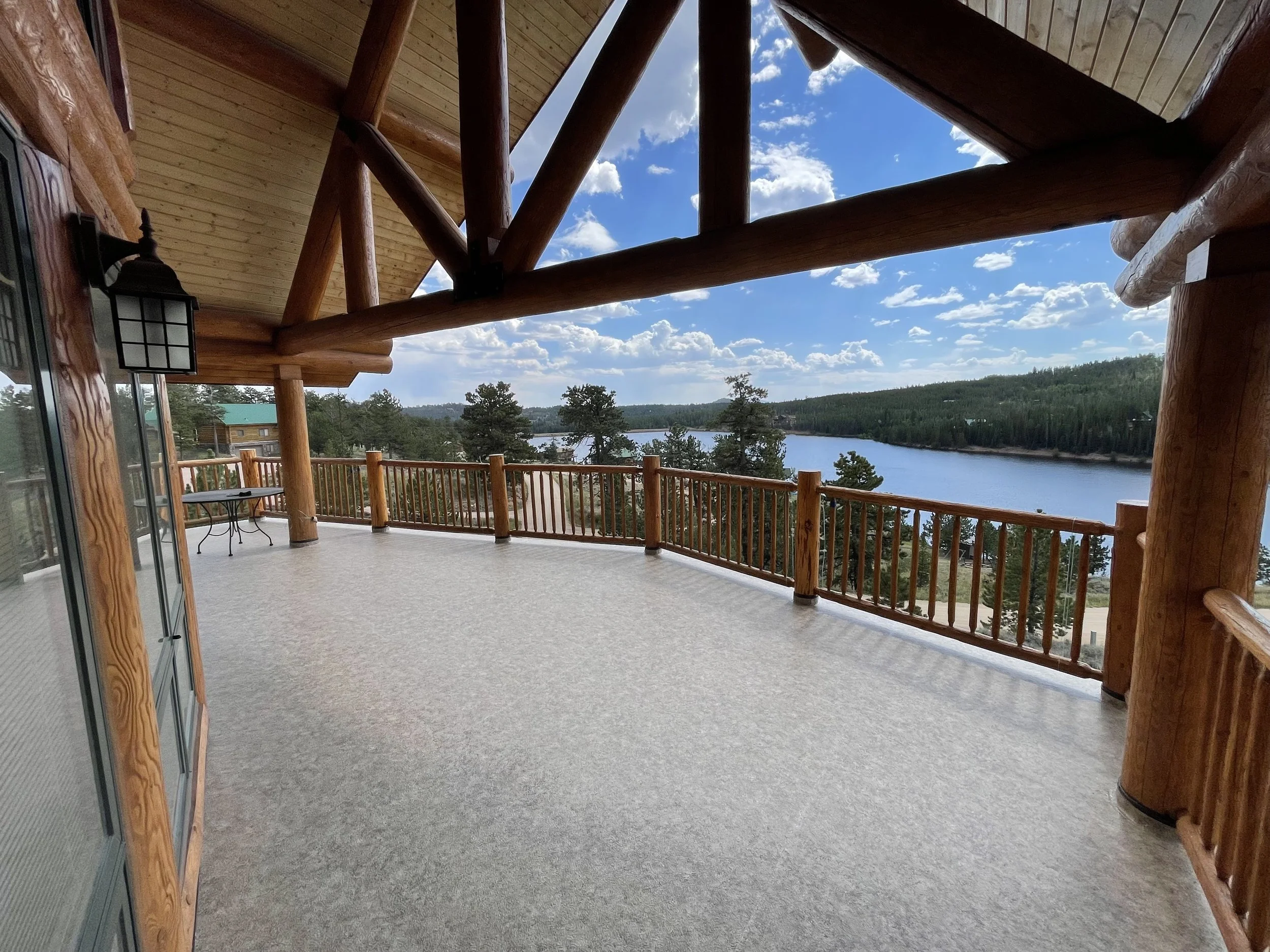 Large outdoor balcony with wooden railings and support beams, overlooking a river and forested landscape under a partly cloudy sky.