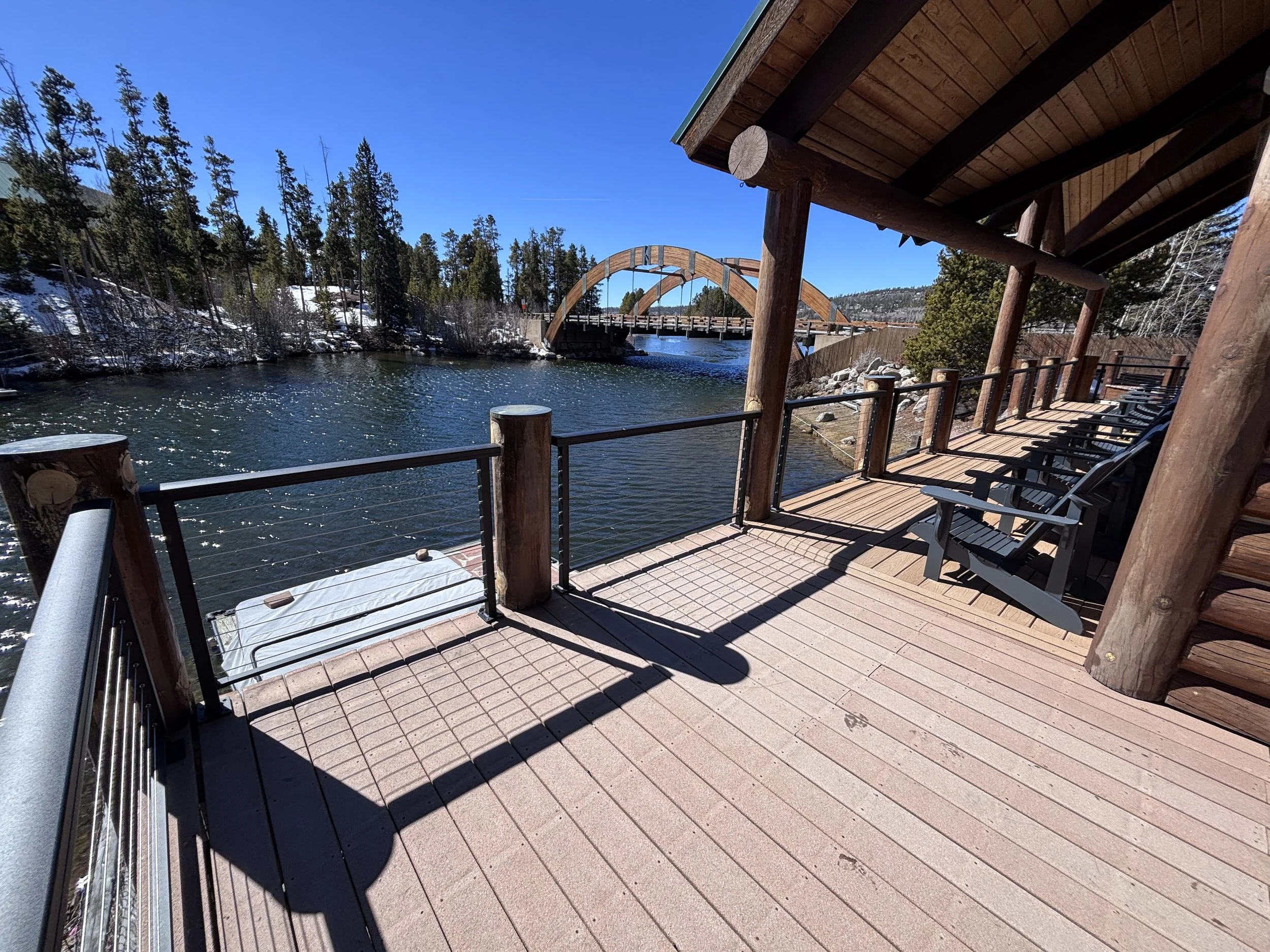 Wooden deck with lounge chairs overlooking a river with a bridge and forested hills in the background, under a clear blue sky.