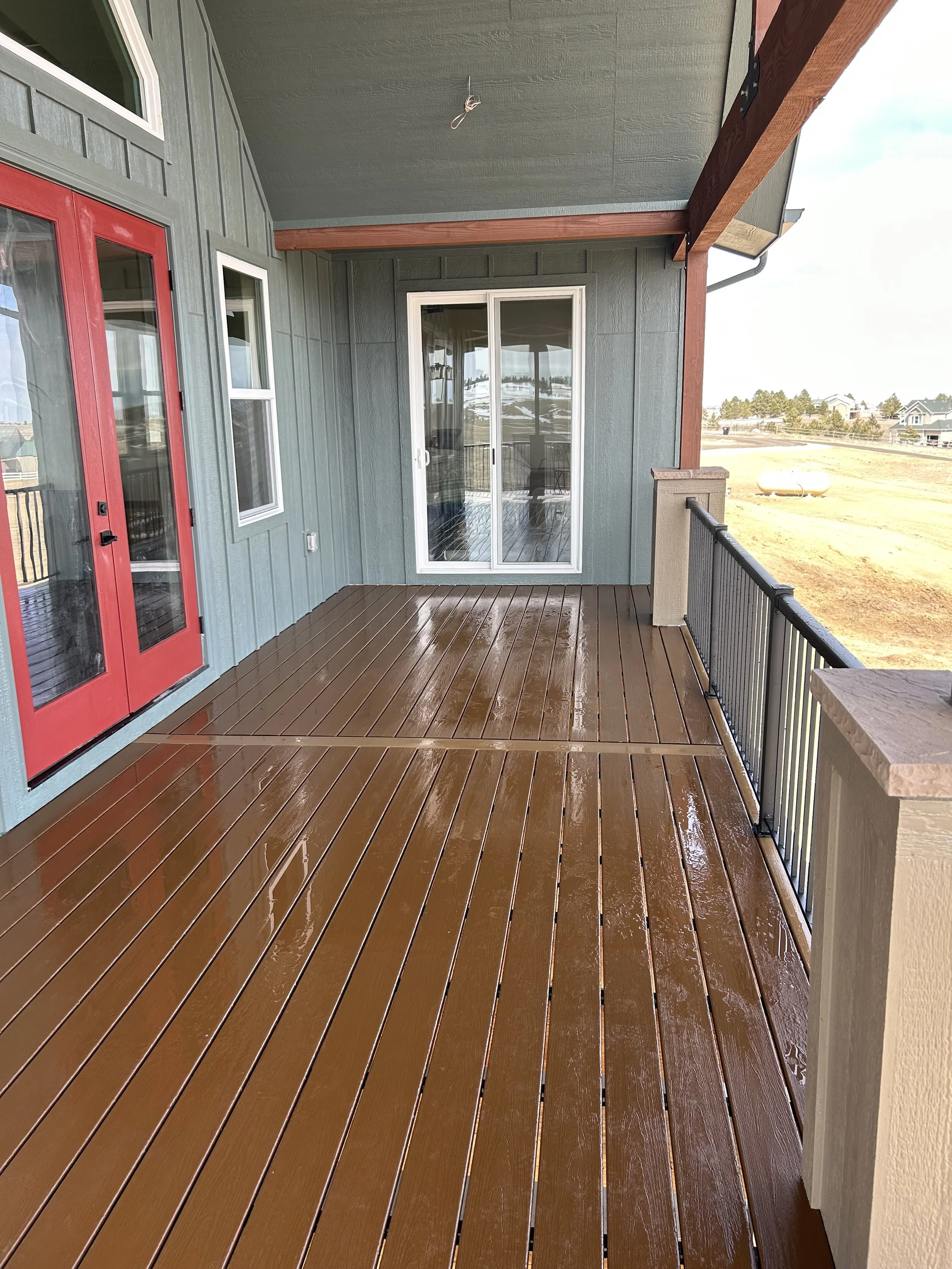 A newly constructed balcony with wet wooden decking, green exterior walls, a red door, and a sliding glass door, overlooking open fields and houses in the distance.