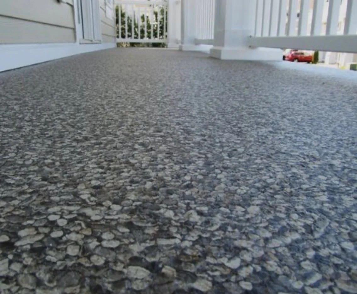 Close-up of a textured gray outdoor balcony floor, with wooden railing and white siding of a house visible in the background.