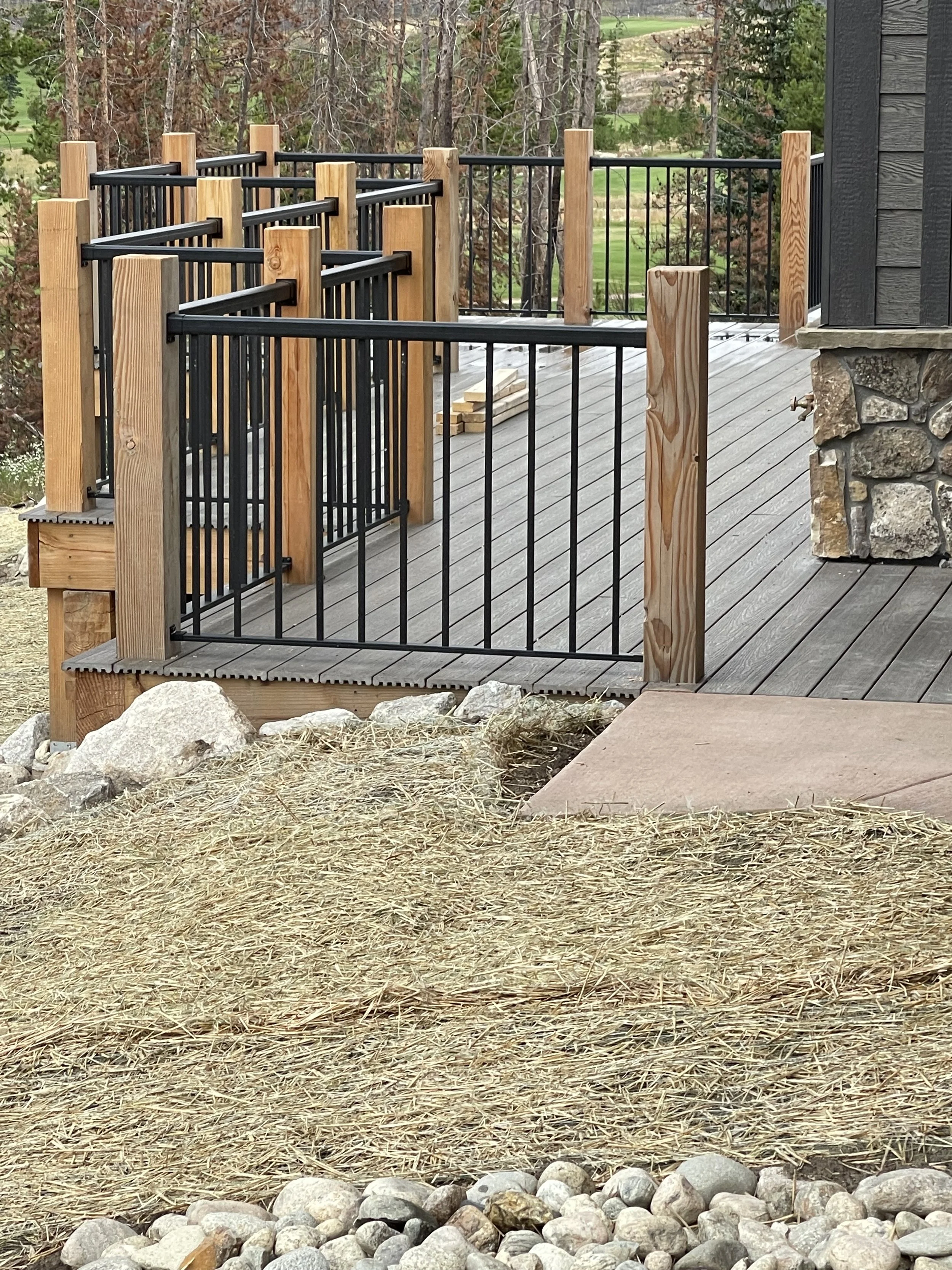 Part of a wooden deck with black metal safety railings, attached to a house with stone foundation, overlooking a landscape with trees and grass.