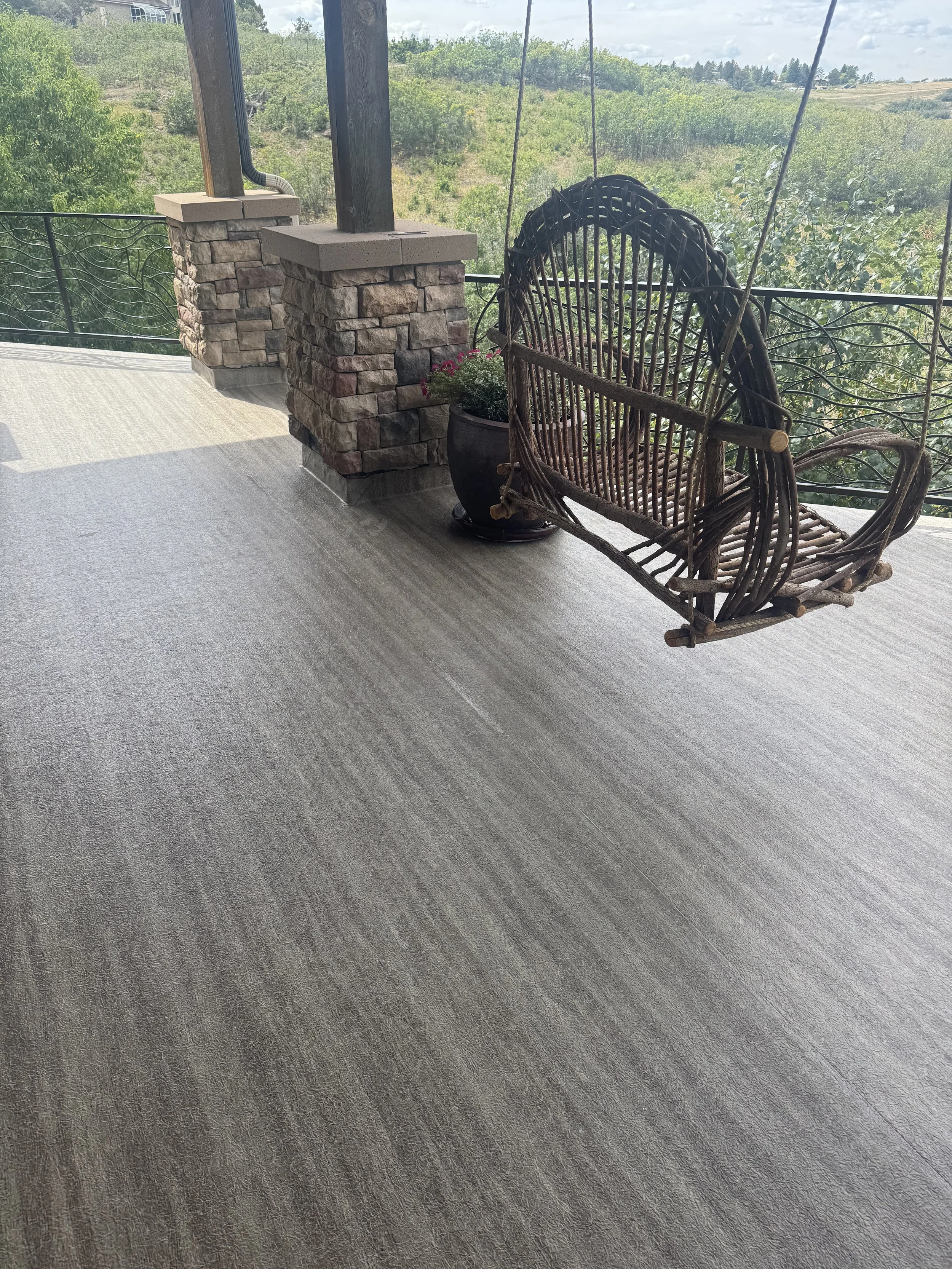 A balcony with a stone pillar, a hanging wicker chair, and a potted plant, overlooking a green landscape and cloudy sky.