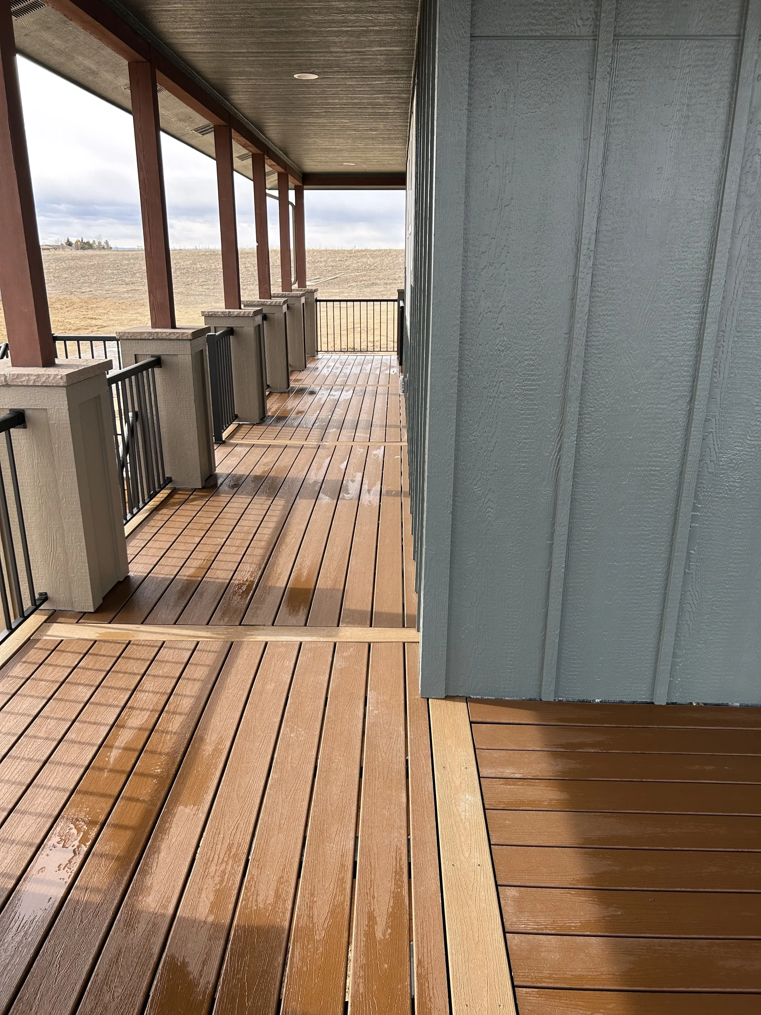 A wooden balcony with wet floorboards, beige pillars, black metal railings, and a blue wall with vertical paneling, overlooking an open field under a cloudy sky.