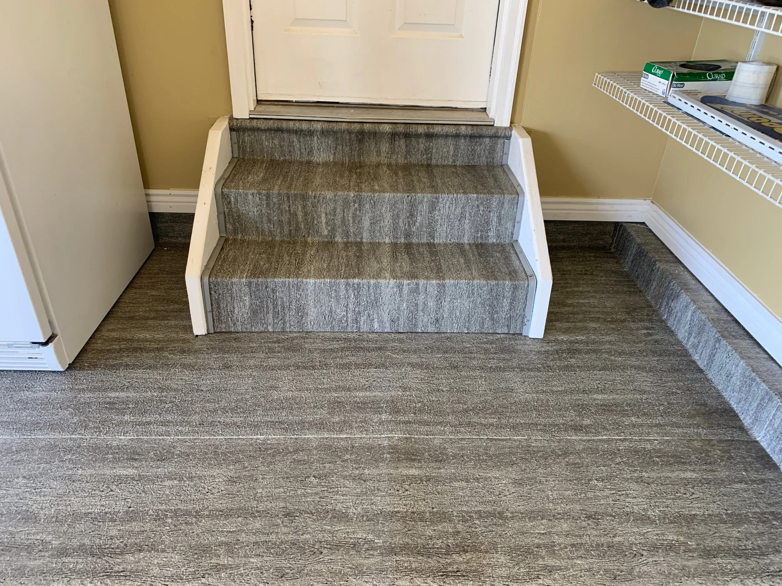 Upgraded carpeted stairs leading to a door, with a laundry shelf on the right and a refrigerator on the left.