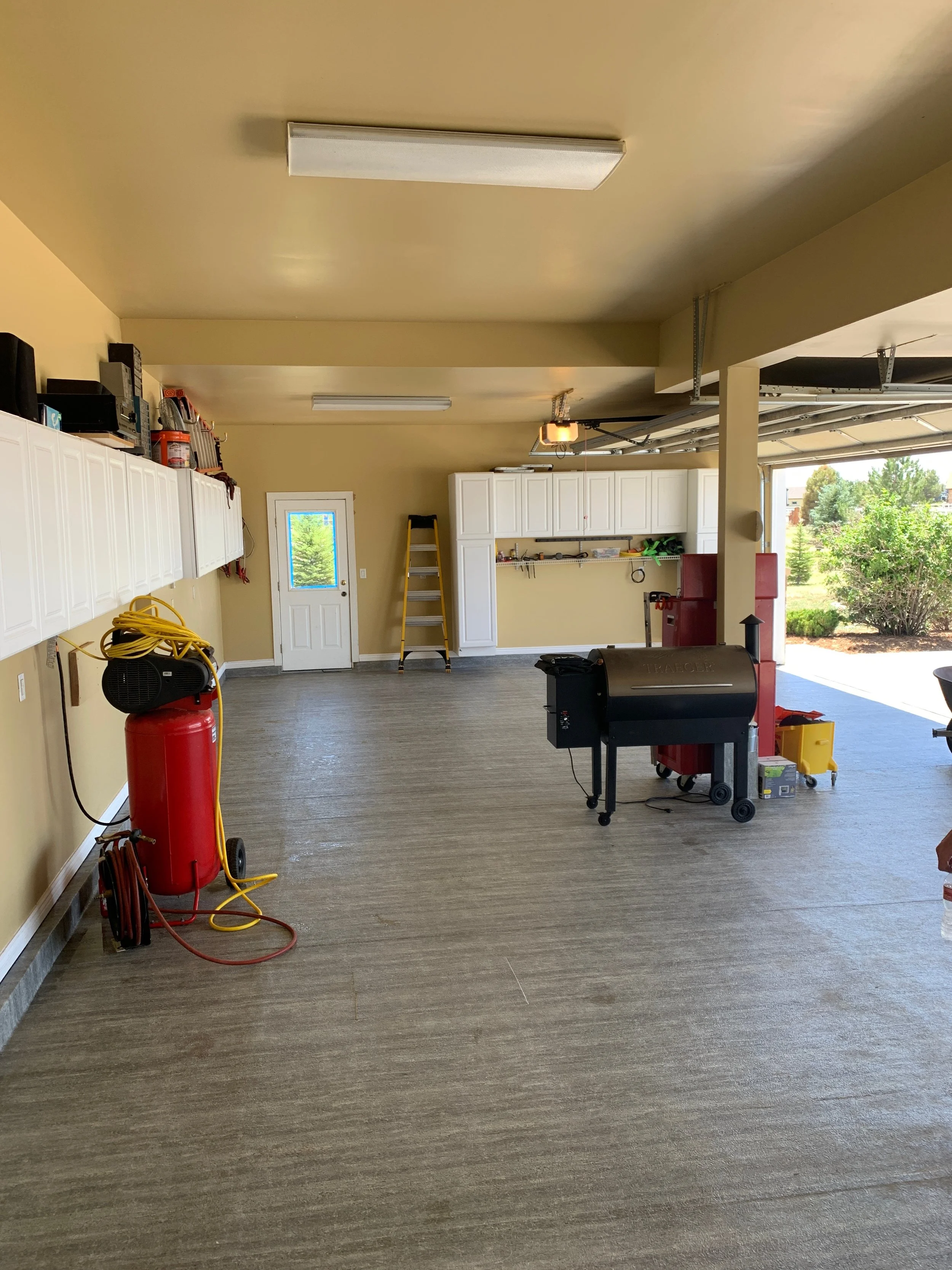 An empty garage with yellow walls, white cabinets along the back wall, a ladder, a black grill, a red fire extinguisher, and tools and storage items. Bright natural light coming from the garage door on the right, with a view of greenery outside.