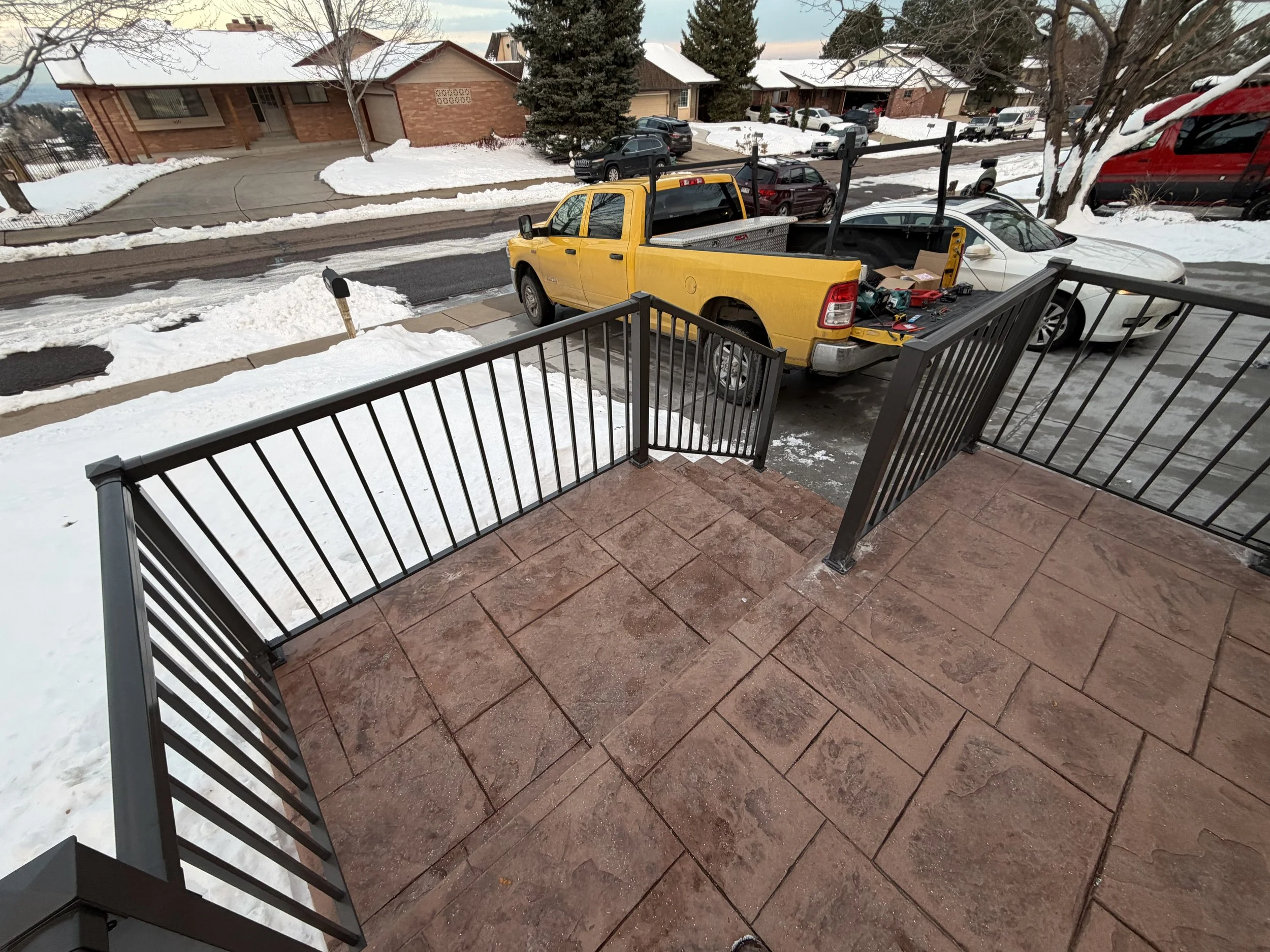 View of a snow-covered front yard with a black metal railing on a brick porch, a yellow pickup truck with tools in its bed, and parked cars on the street in a residential neighborhood during winter.