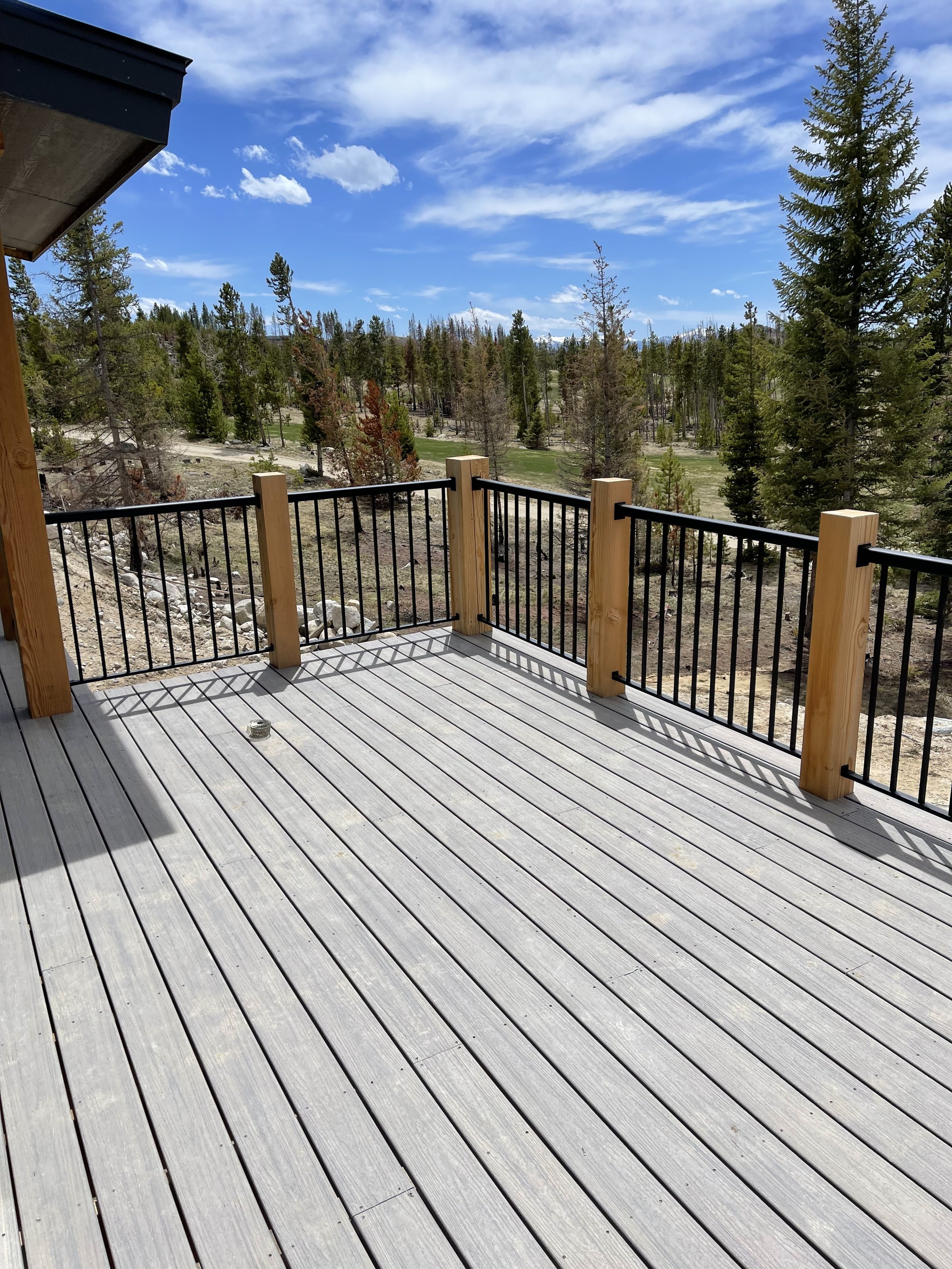 View from a new wooden deck with black metal railing, overlooking a landscape of tall evergreen trees, grassy areas, and distant mountains under a partly cloudy blue sky.