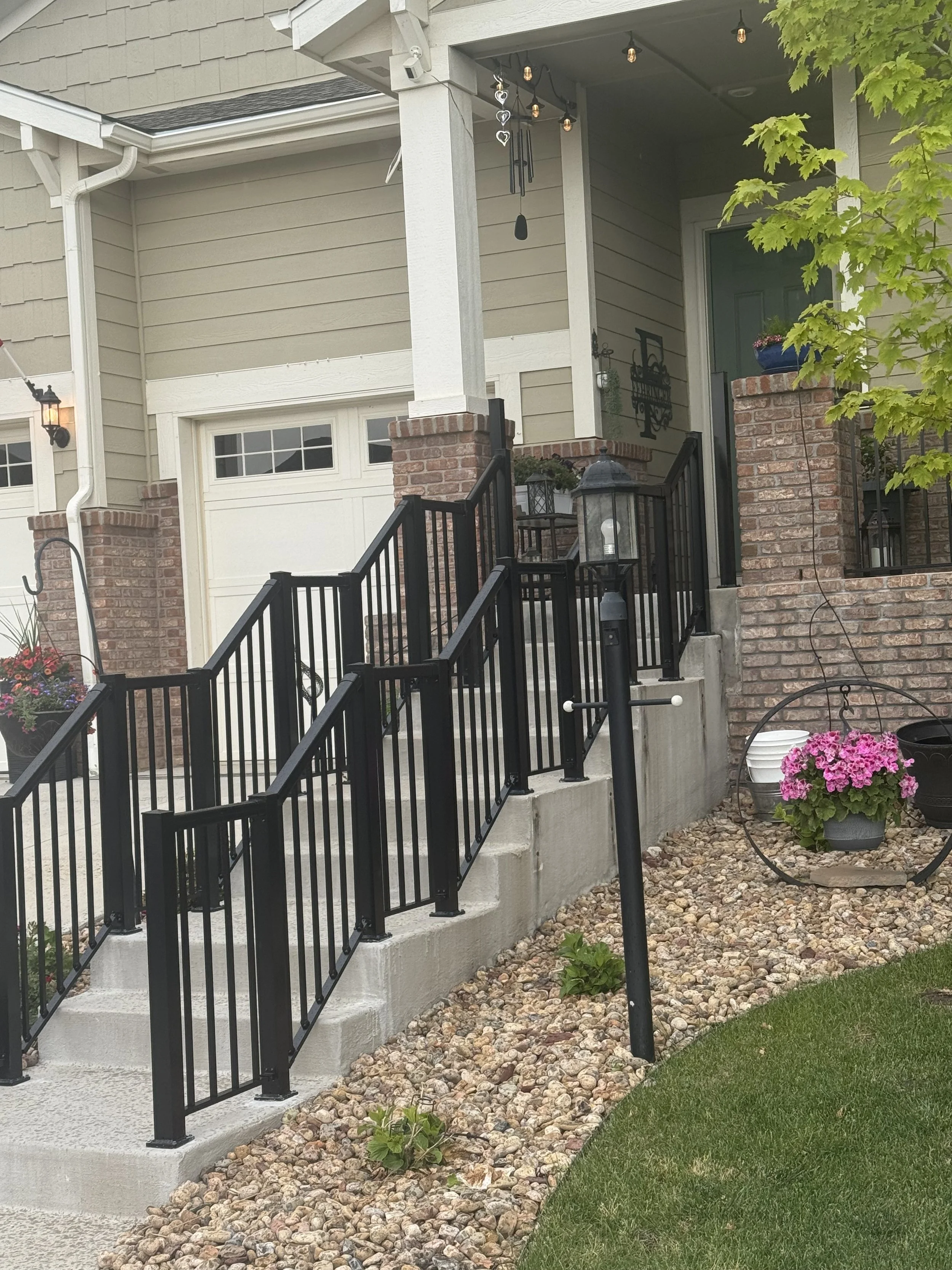 Front porch stairs with black metal railings leading to a light gray front door, decorated with potted pink and purple flowers, brick pillars, and string lights hanging from the porch ceiling. A tree branch with green leaves partially extends into th
