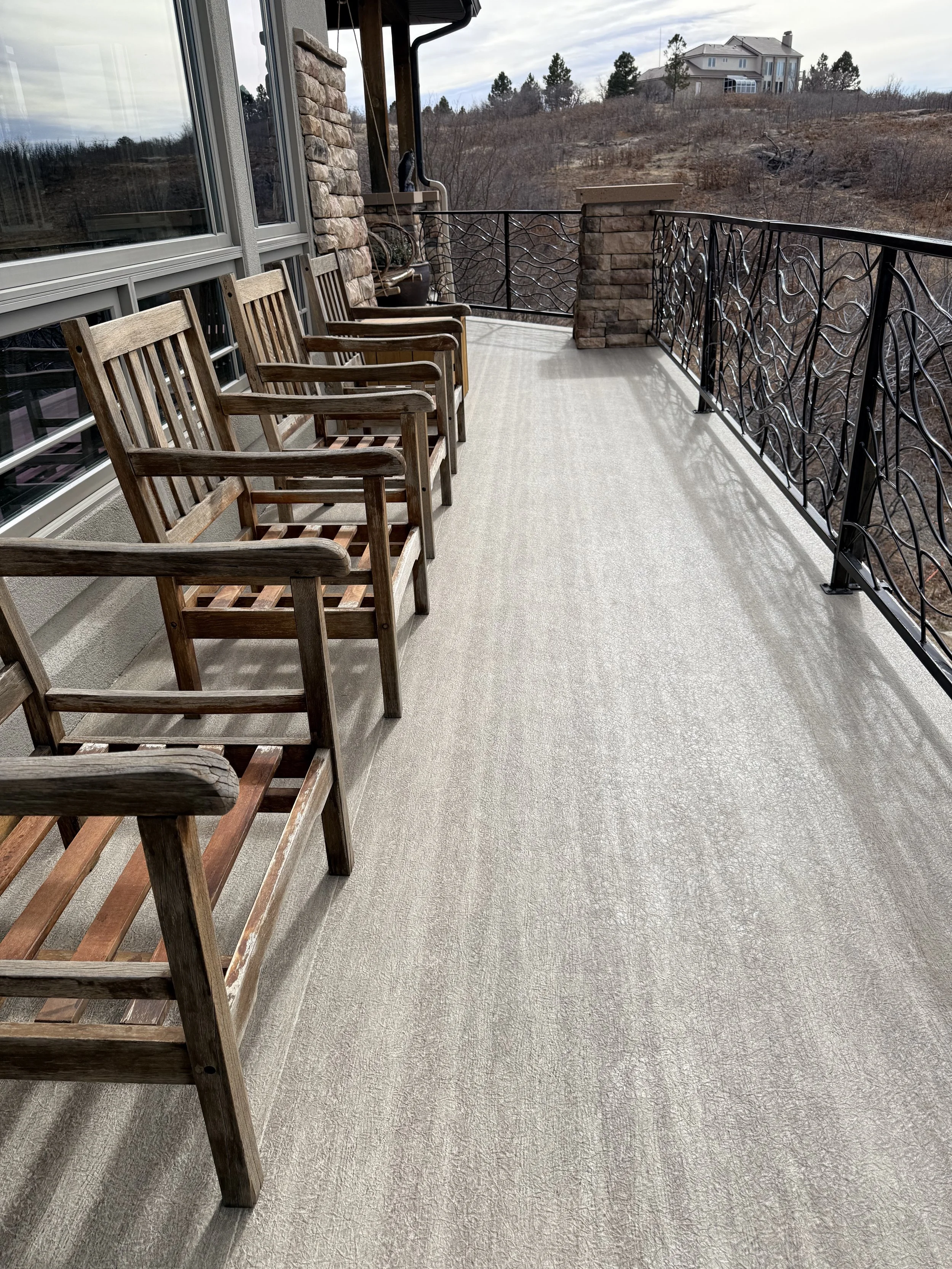 Empty outdoor balcony with four wooden chairs, decorative black metal railing, stone wall, and scenic view of hilly landscape with houses and bare trees.