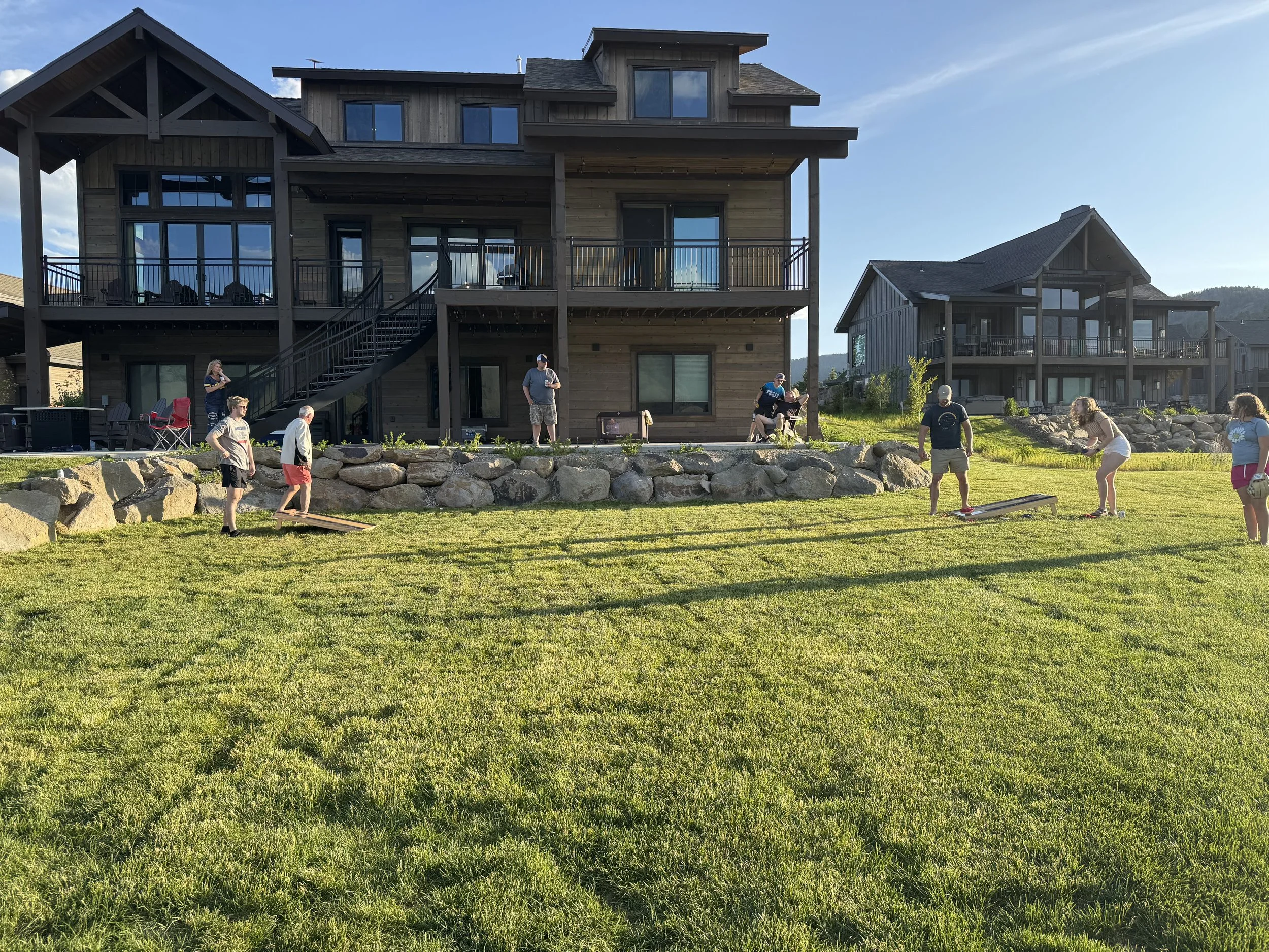 People playing bean bag toss on the lawn outside of large modern houses in a residential neighborhood on a sunny day.