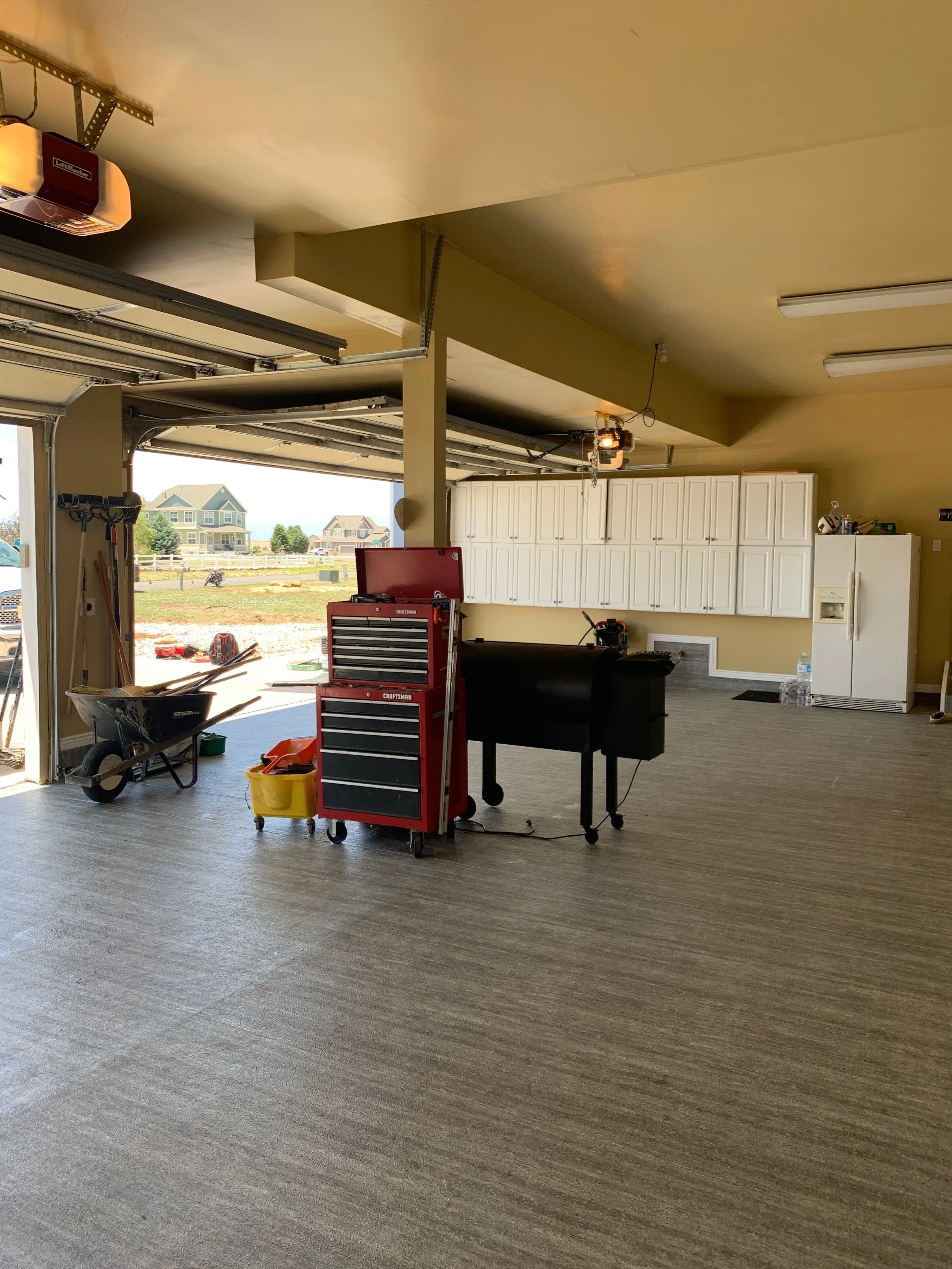 Empty garage with tools, a wheelbarrow, and a grill inside, and houses visible through the open garage door.