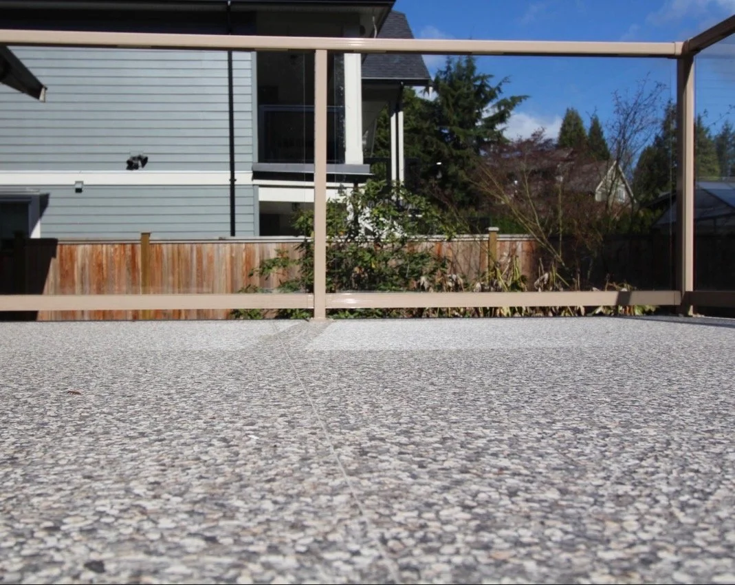 A view of a backyard patio with a soapstone or concrete surface, wooden and glass railing, and residential houses with trees in the background under a partly cloudy blue sky.