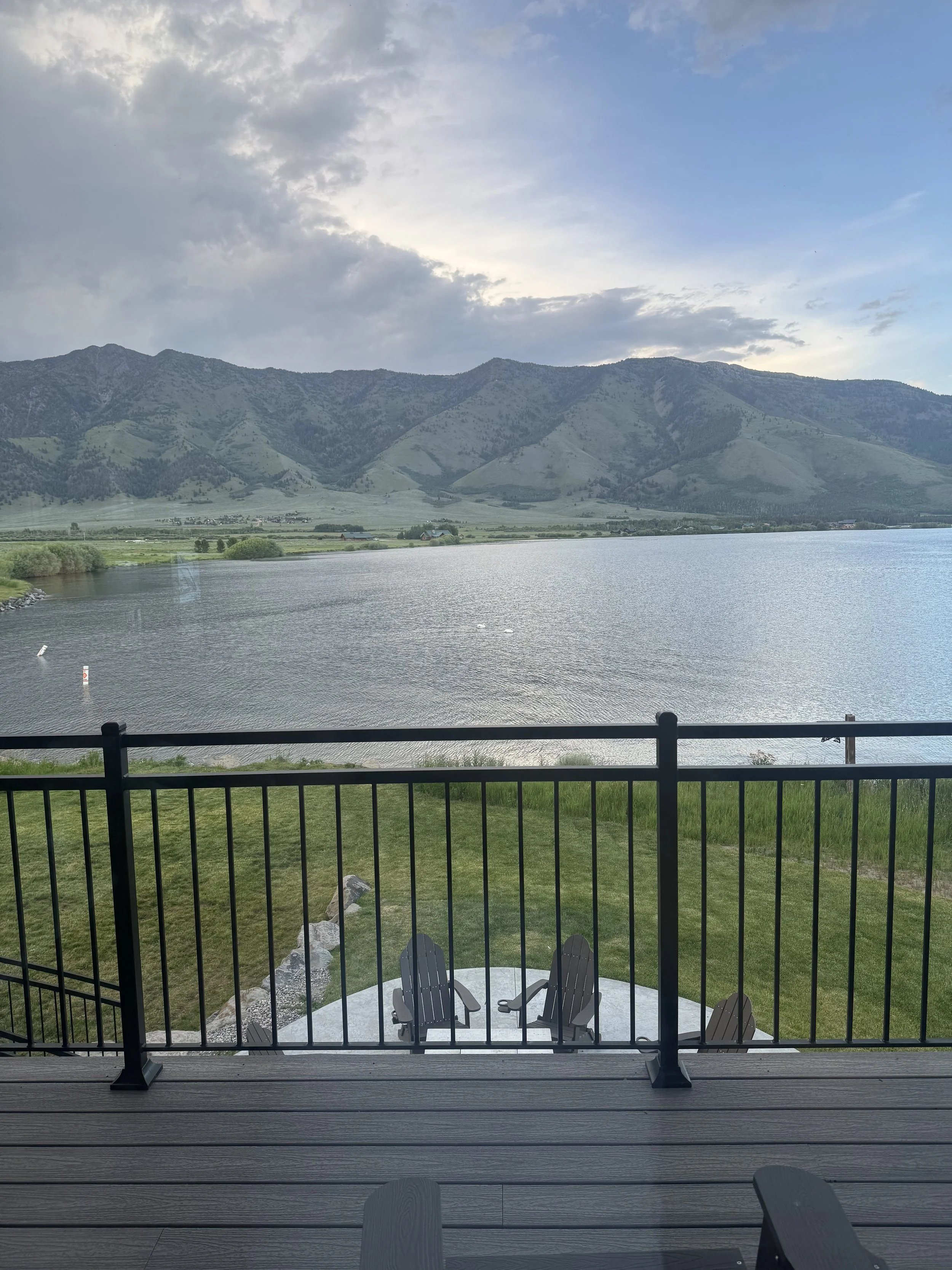 View from a balcony with black railing, overlooking a lake, green grass, Adirondack chairs, and distant mountains under a cloudy sky.