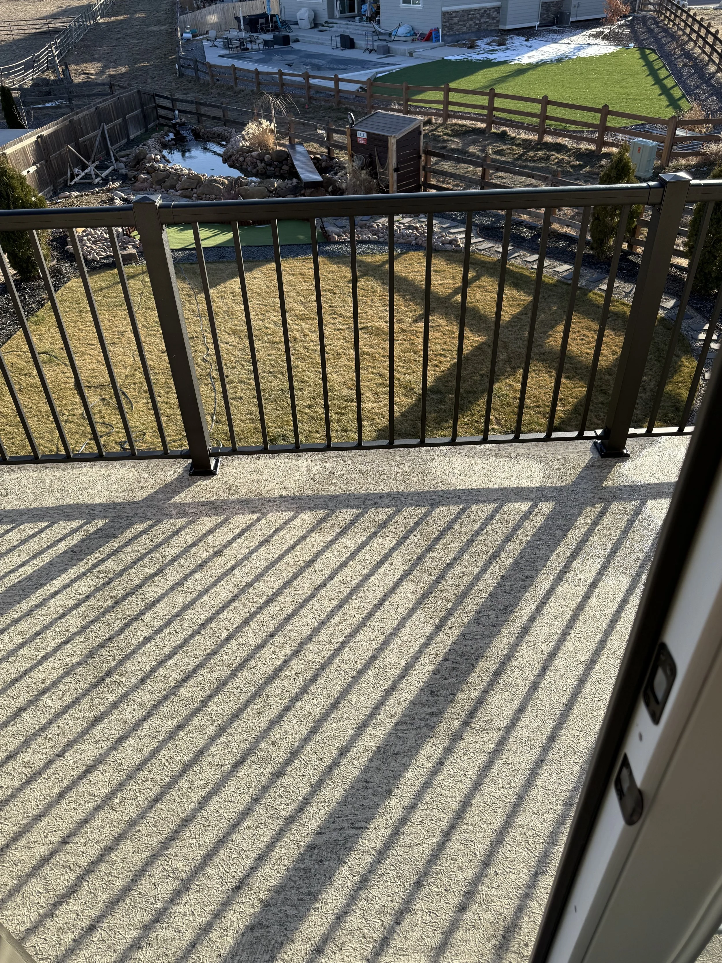 View from a balcony with a black metal railing casting shadows on a concrete floor, overlooking a backyard with a lawn, a small pond, and a fenced area.