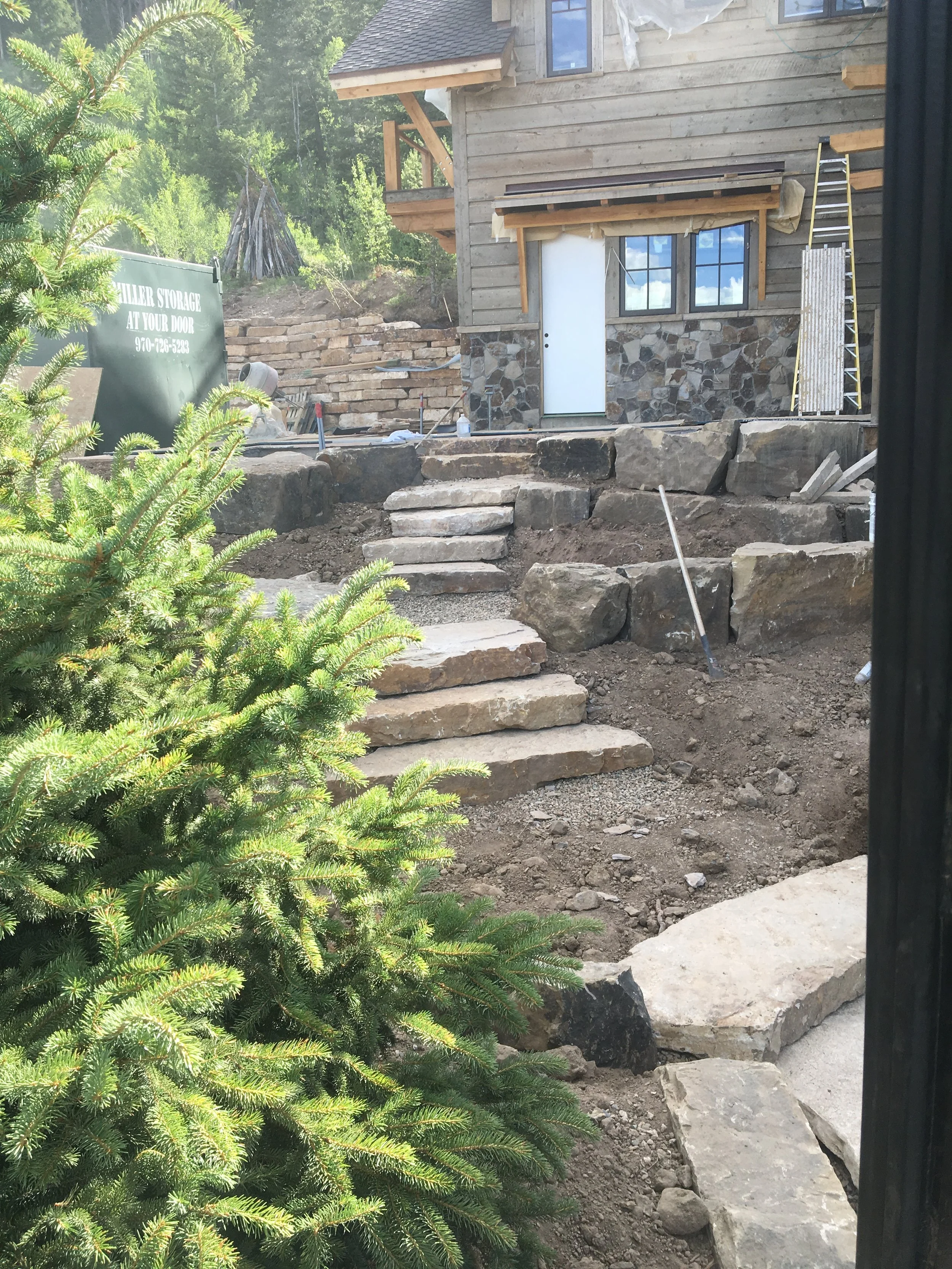 Construction site of a stone staircase leading to a house with wooden exterior siding. There are tools and building materials around, and a ladder leaning against the house. A pine tree is in the foreground.