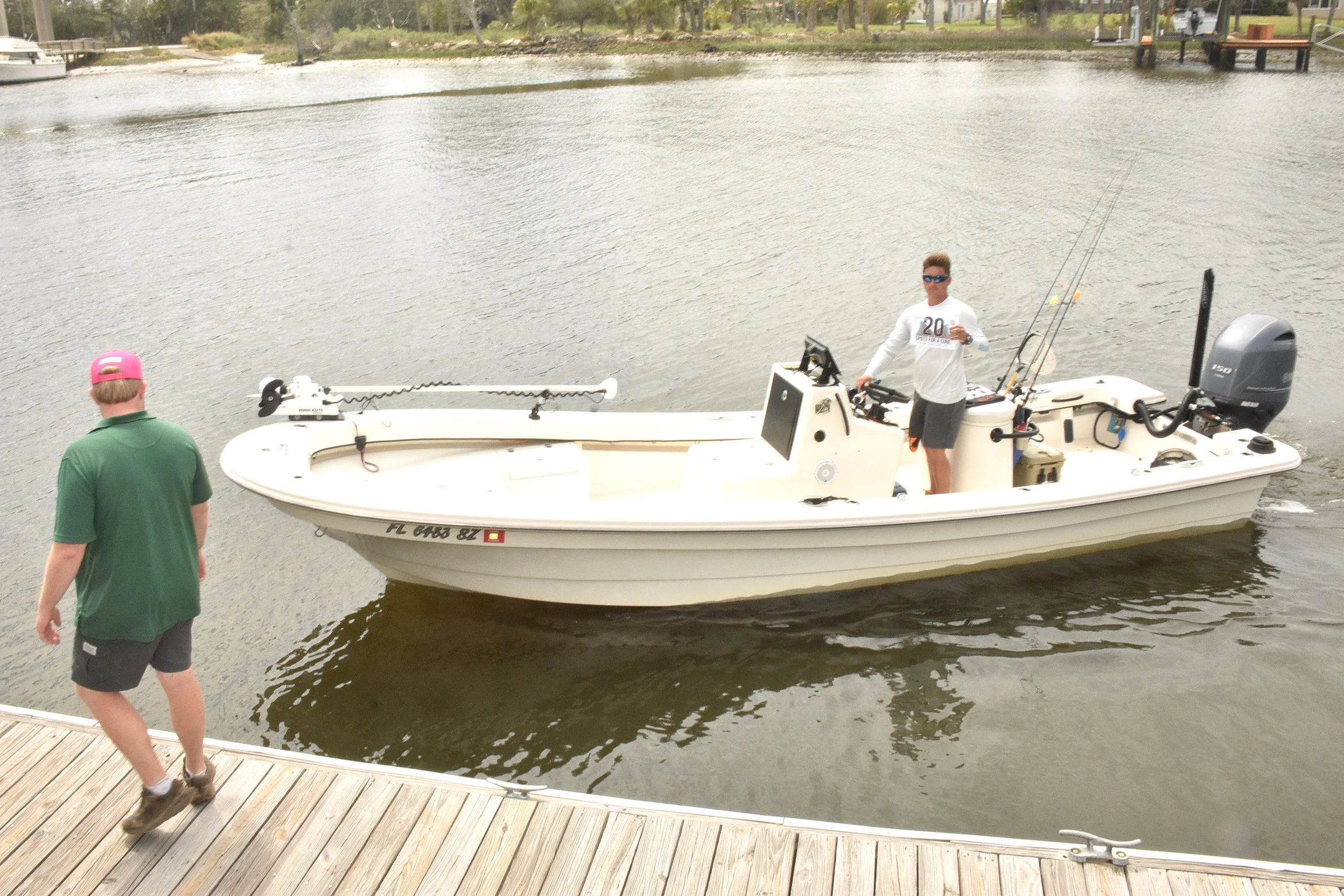 A small white fishing boat with two men, one standing on the boat holding fishing rods and the other walking on the dock wearing a green shirt and pink cap. The boat is on a body of water with a wooden dock in the foreground.
