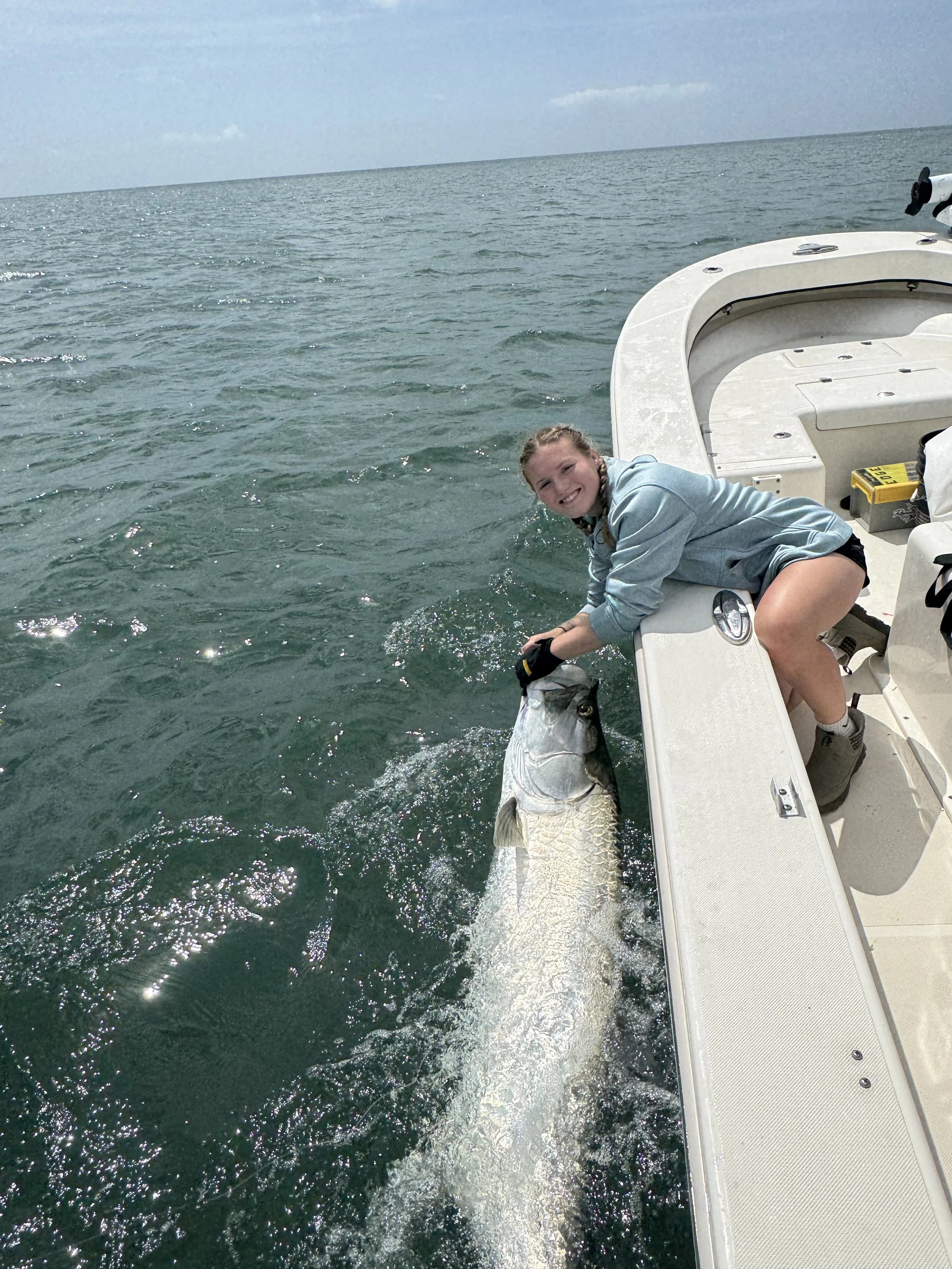 A girl on a boat holding a large tarpon she caught, with the open water in the background.