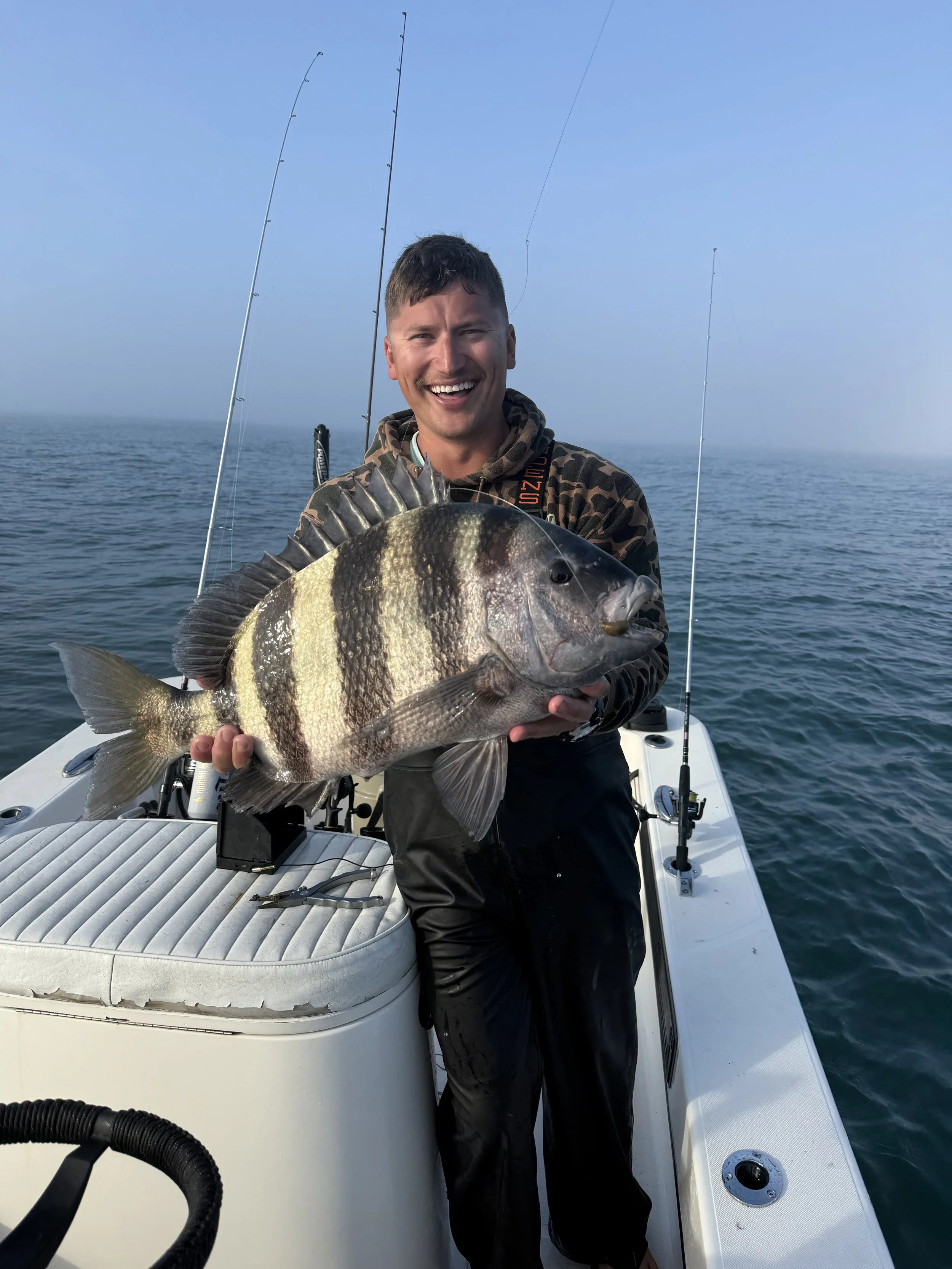 A man on a boat holding a large sheephead, smiling at the camera, with fishing poles in the background, on a calm sea during daytime.