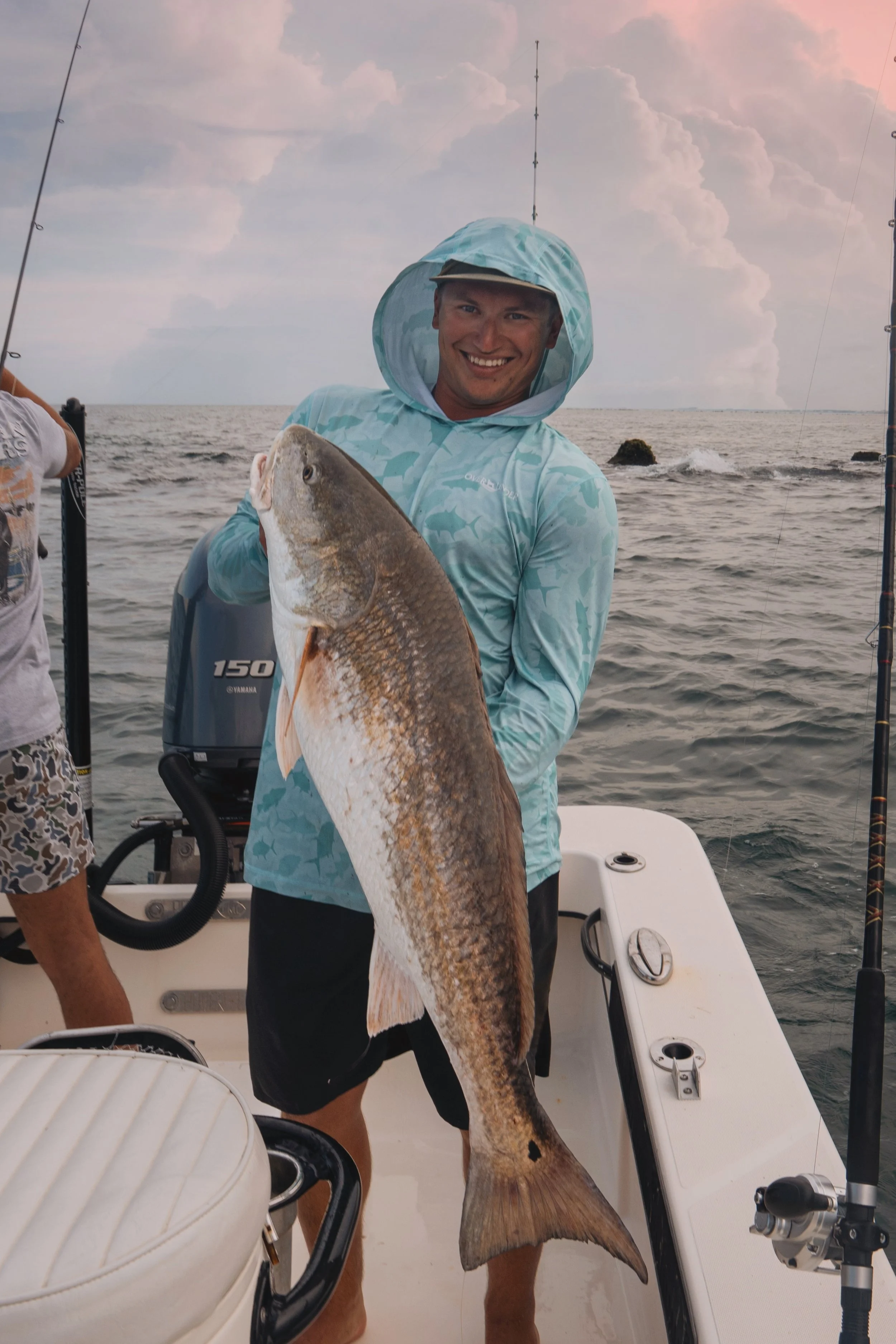 A man wearing a blue hoodie holding a large redfish on a boat at sea during sunset.