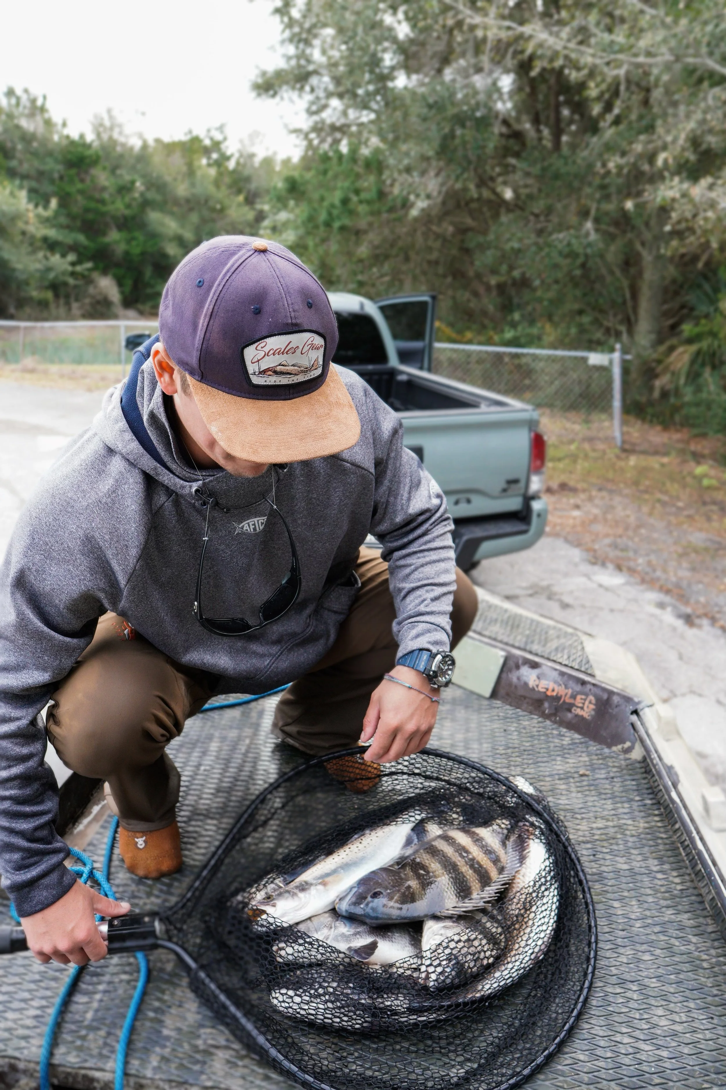 Person in gray hoodie squatting on a boat deck, holding a fishing net with several caught fish, with a pickup truck and trees in the background.