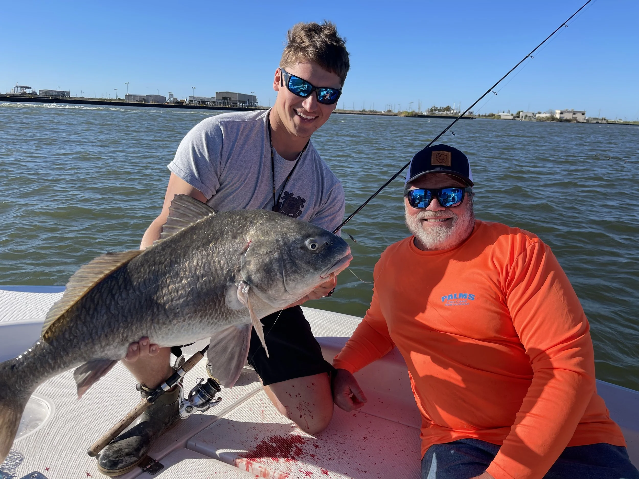 Two men on a boat holding a large fish, smiling with water in the background.