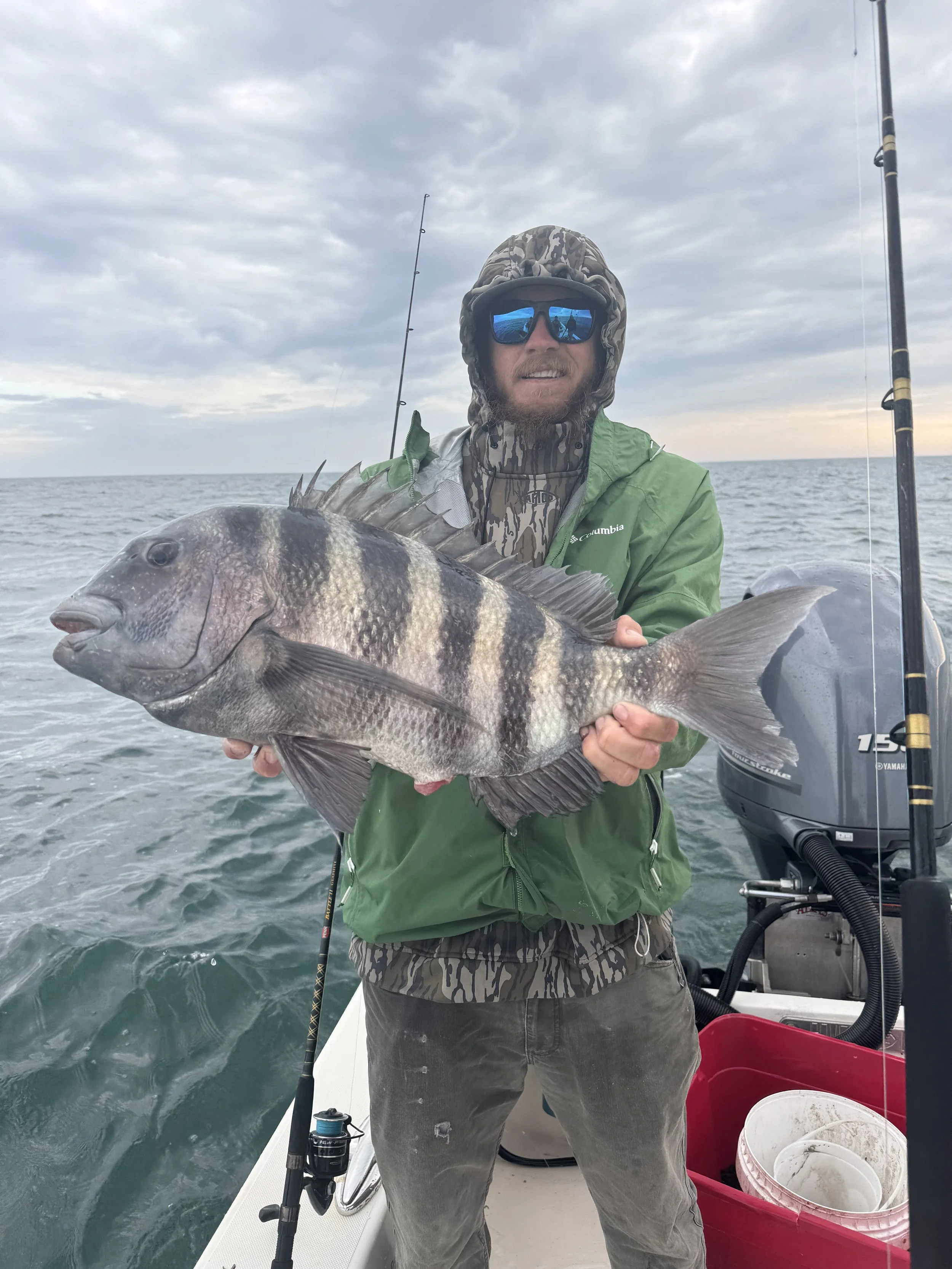 A man wearing a green jacket, camo hood, and sunglasses on a boat, holding a large sheephead he caught, with the ocean and cloudy sky in the background.