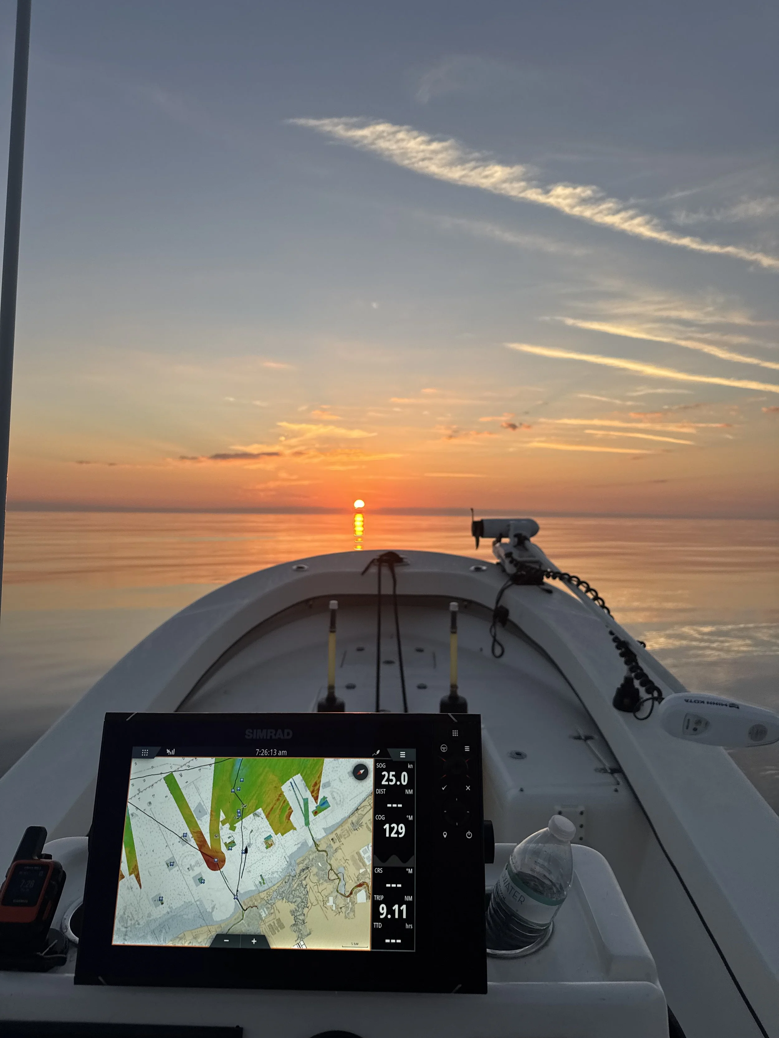 View from a boat at sunset, with navigational equipment and a water bottle on deck, looking towards the calm water and a colorful sky with clouds.