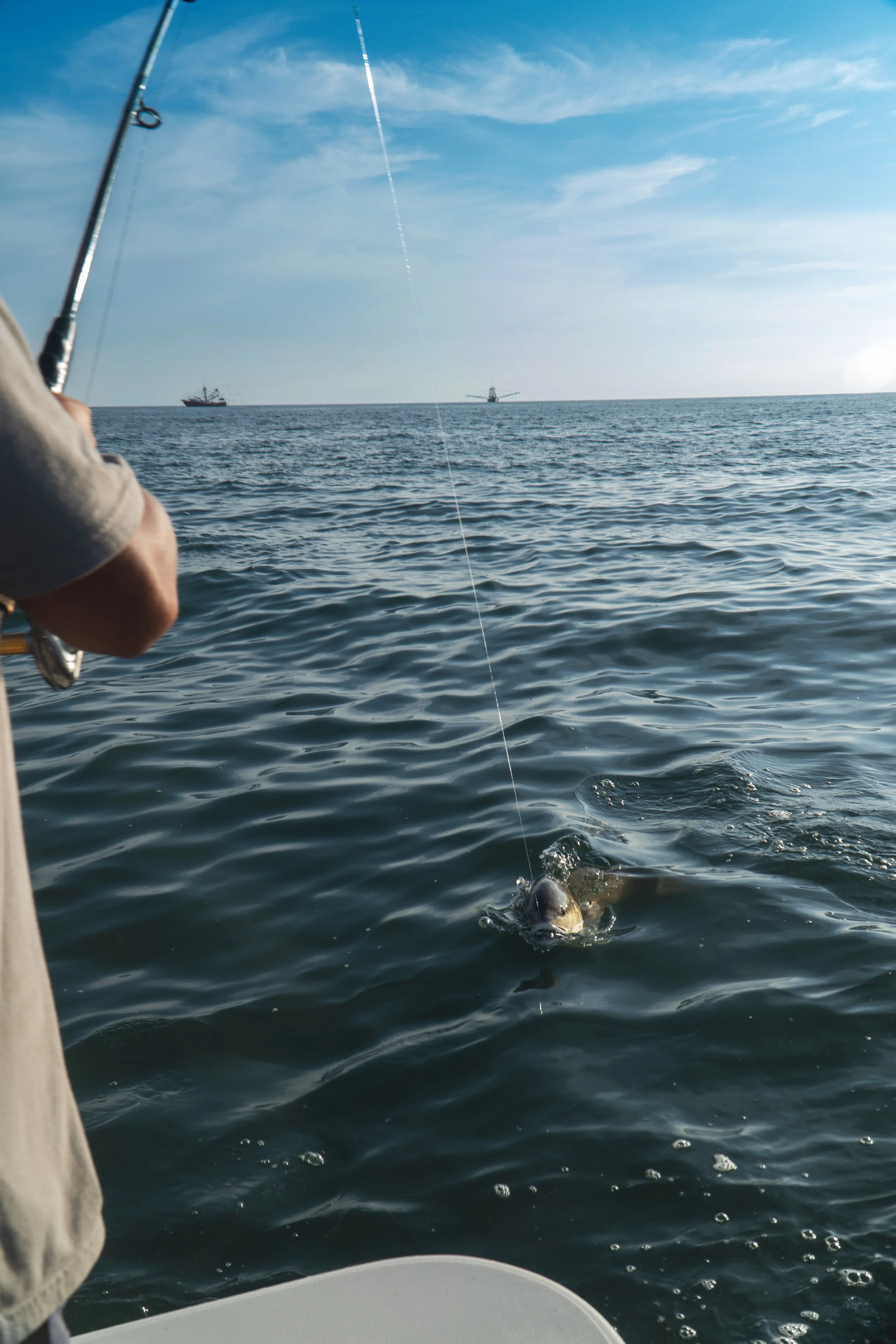 Person fishing on a boat in the ocean, with a redfish caught on the line near the water surface. In the background, there are shrimp boats on the horizon and a partly cloudy sky.