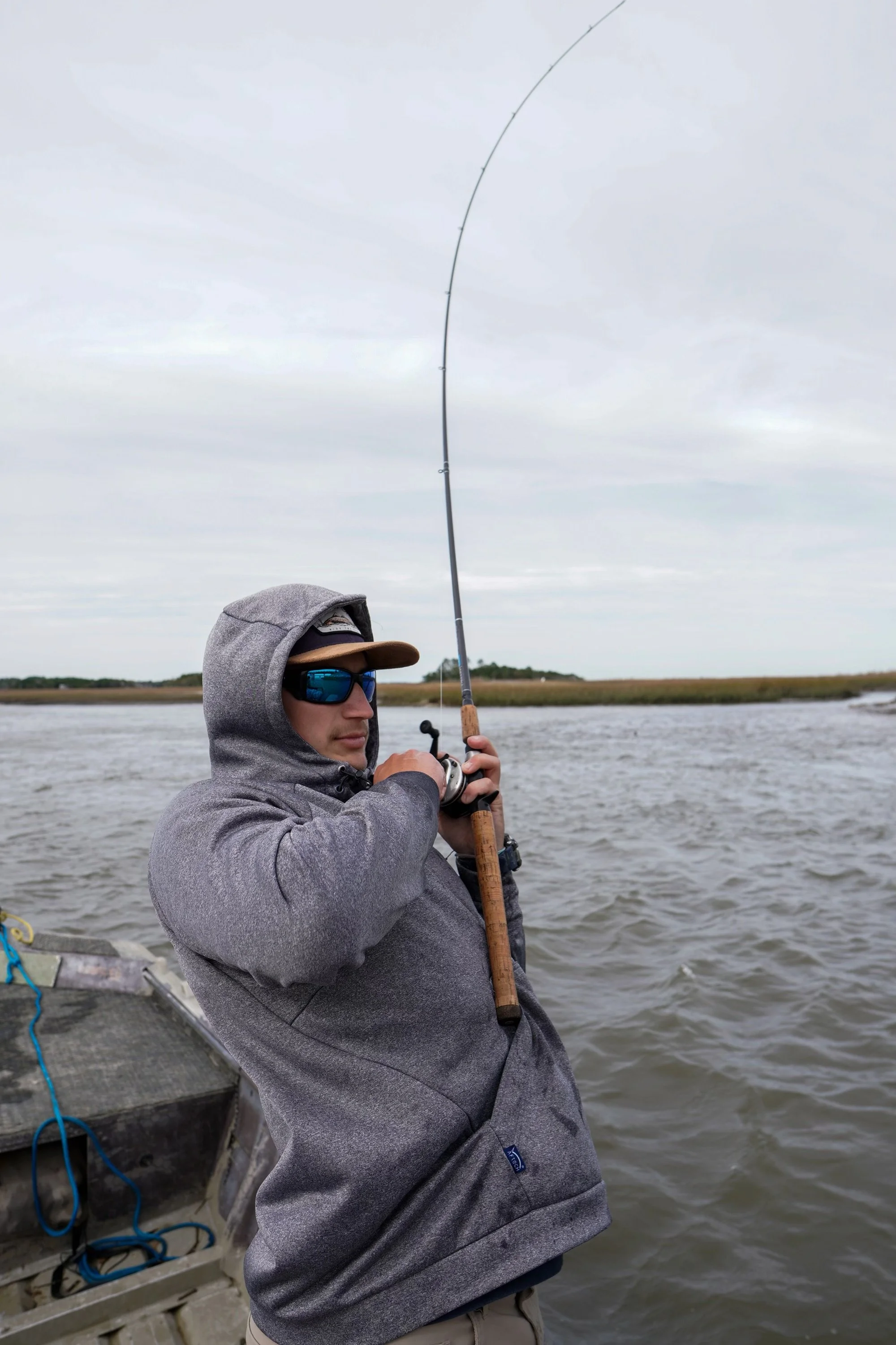 A person wearing sunglasses, a gray hoodie, and a cap is fishing on a boat in a body of water with a cloudy sky overhead.