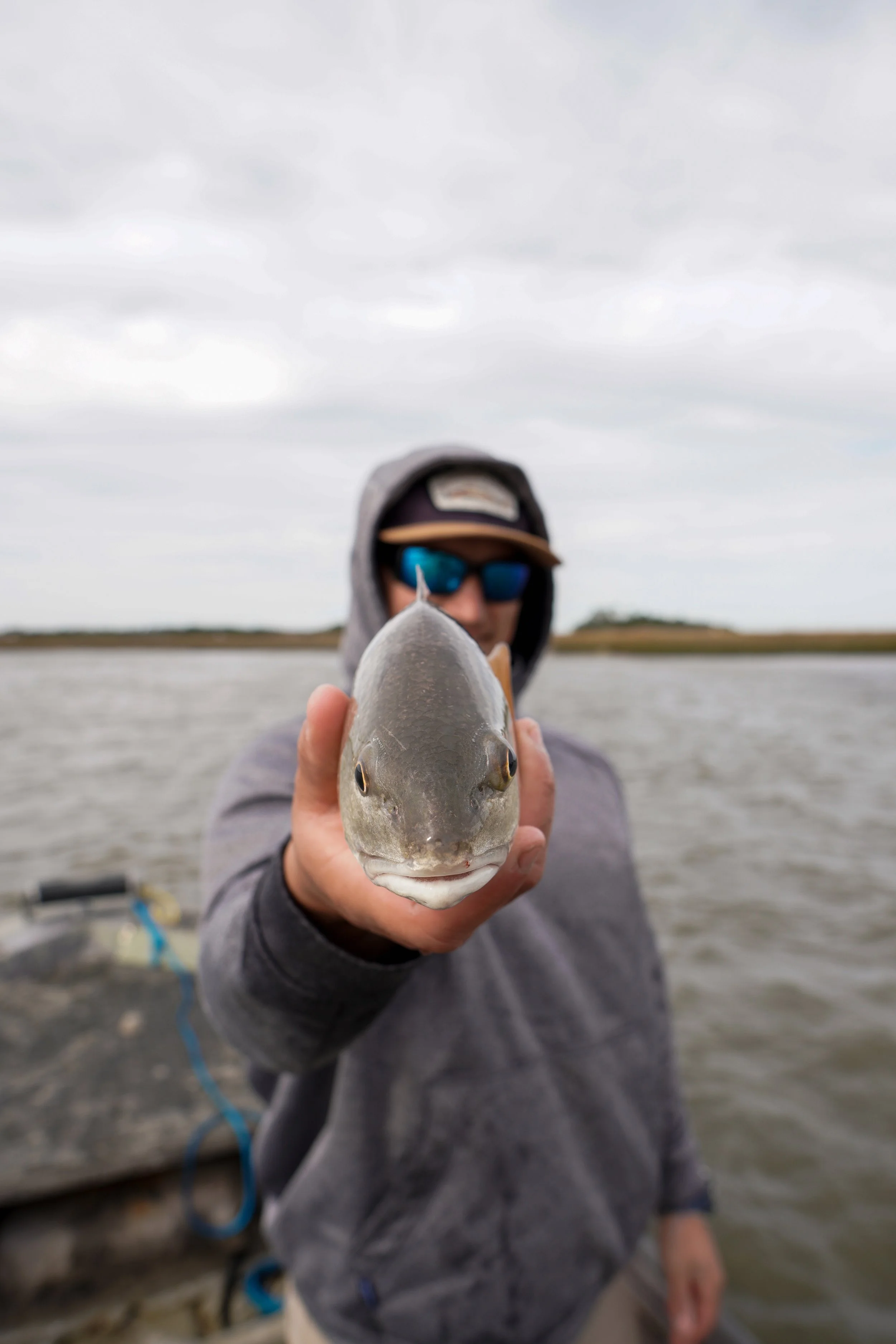 A person wearing sunglasses and a hoodie holding a redfish towards the camera, with a body of water and a cloudy sky in the background.