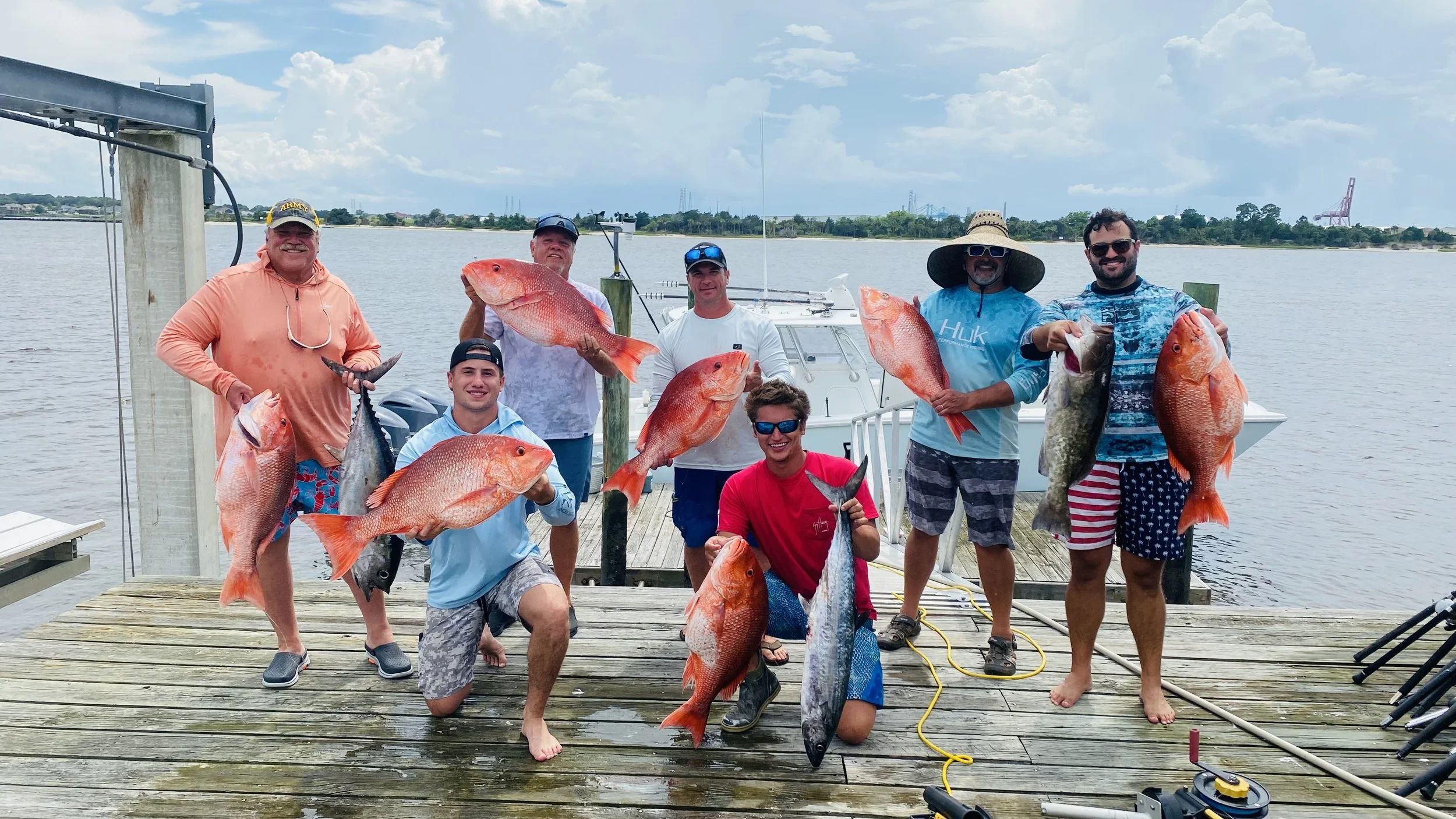 Group of nine men on a dockby holding large red snapper and other fish they caught during a fishing trip by the water, with a boat and cloudy sky in the background.
