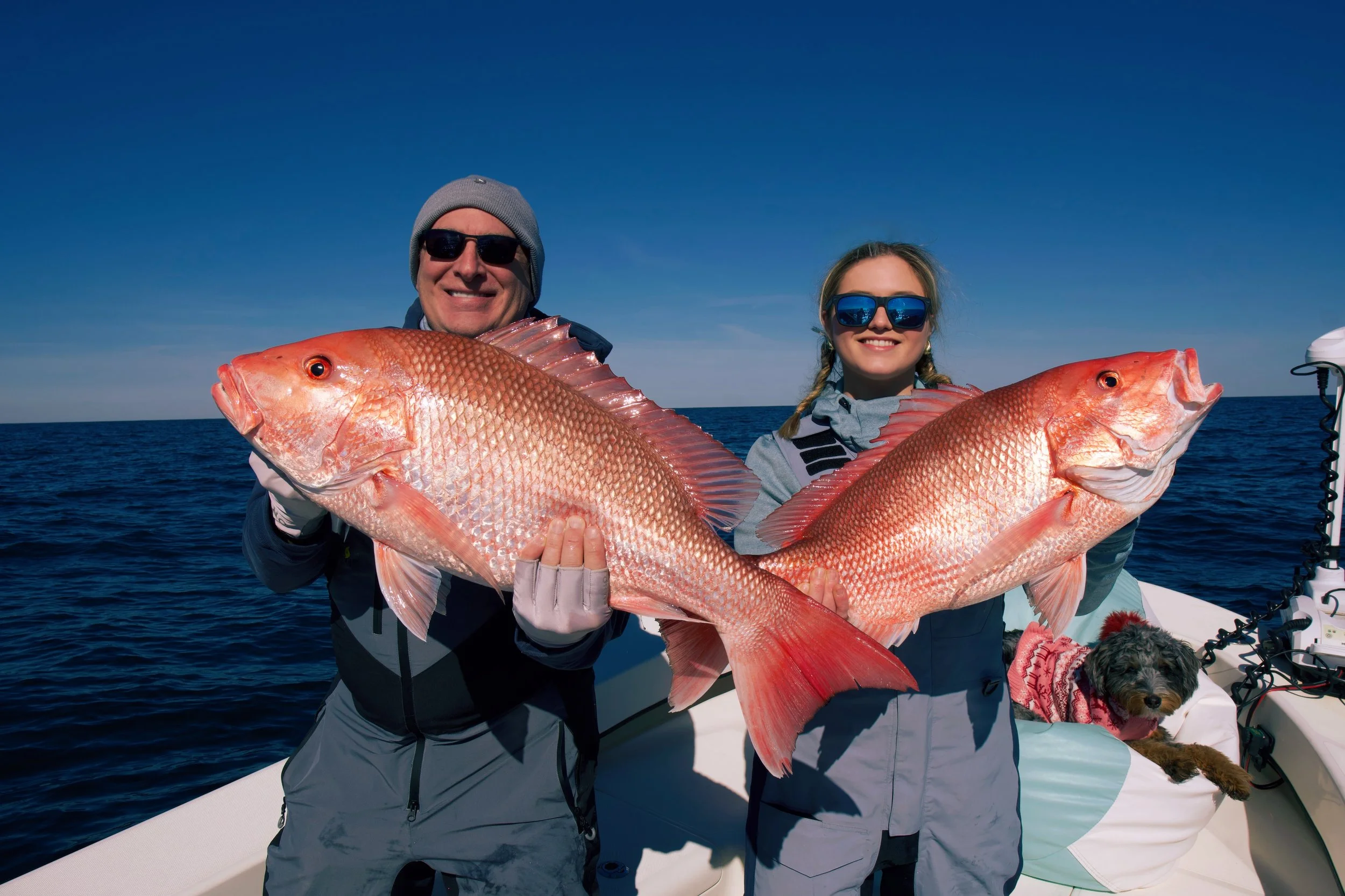 Two people on a boat holding large red fish, with a dog sitting nearby, in the ocean under a clear blue sky.