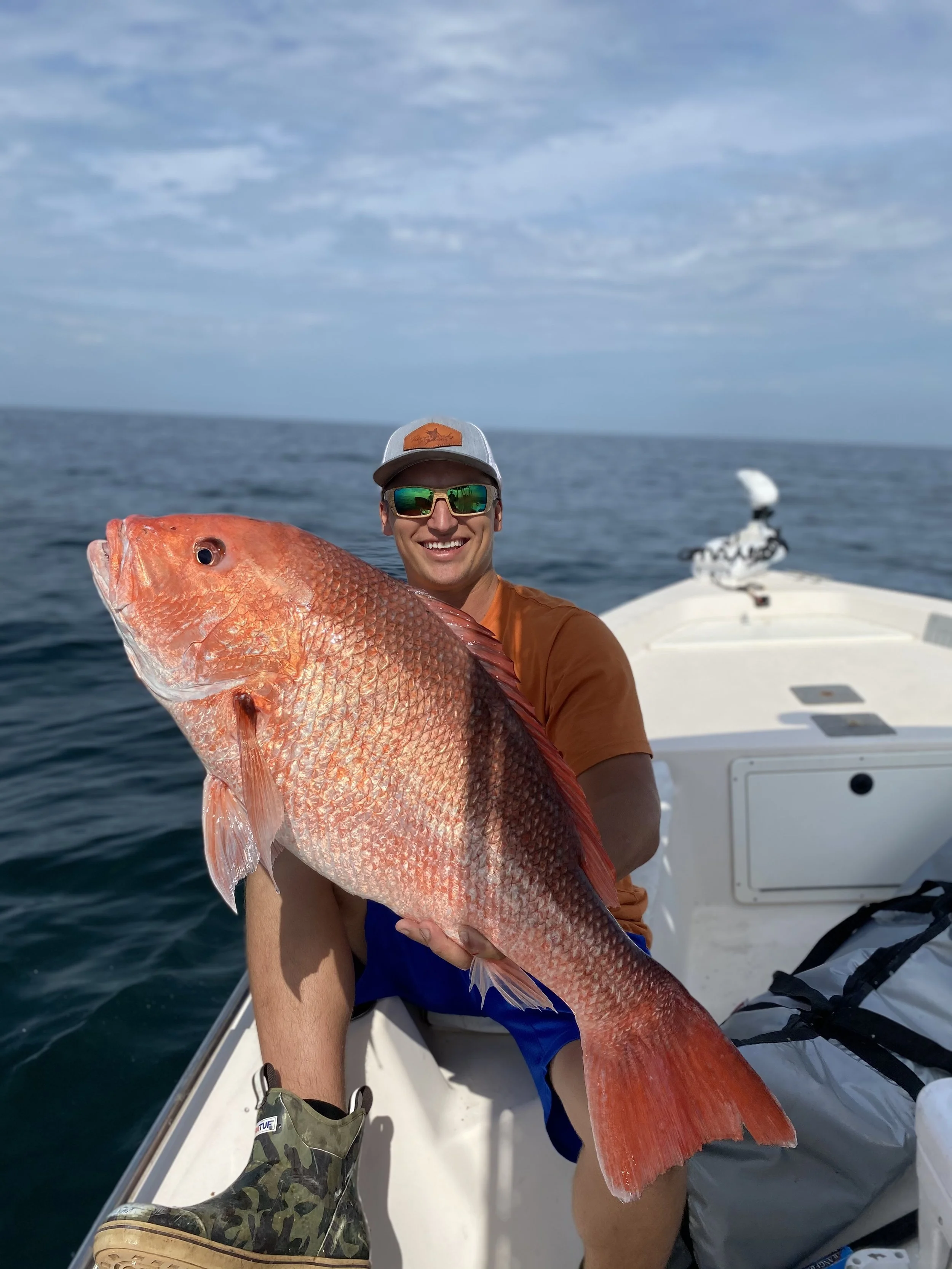 Man wearing sunglasses, holding a large red snapper on a boat in the ocean under a partly cloudy sky.