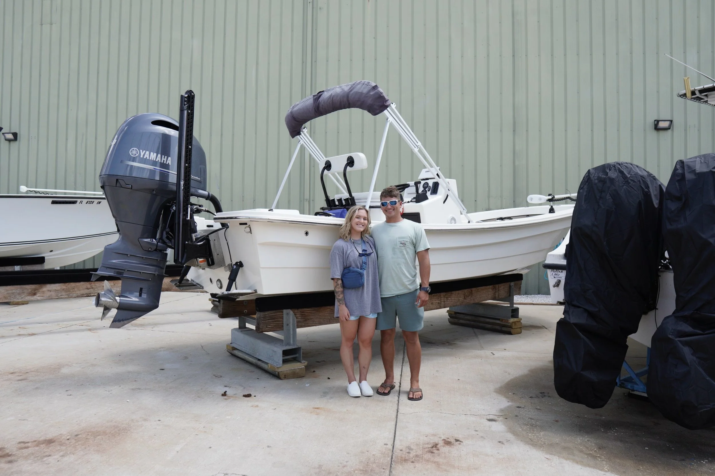 Two people, a woman and a man, standing in front of a white panga with a Yamaha outboard motor on a boat stand outdoors.